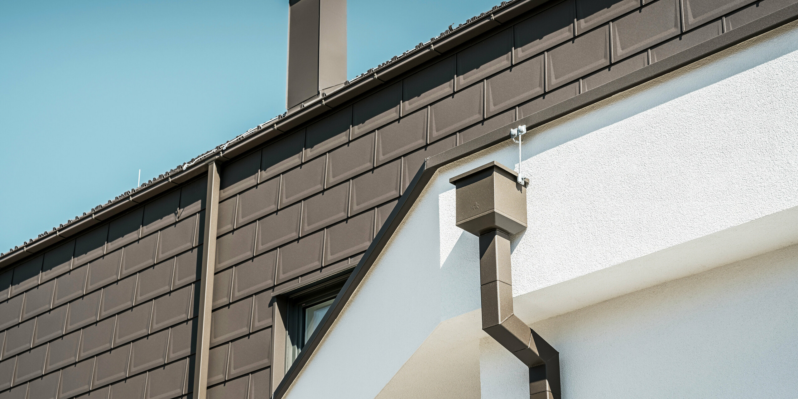Detailaufnahme der Dachkante bzw. der Fassade eines Einfamilienhauses in Neukirchen, Österreich, das mit PREFA Dachplatten R.16 in P.10 Braun ausgestattet ist. Sowohl die Dachplatten, die bei diesem Gebäude als Dach- und Fassadenverkleidung fungieren, als auch das Dachentwässerungssystem, bestehend aus dem Quadratrohr, dem Wasserfangkasten und der Kastenrinne, sind in der gleichen Farbgebung gehalten und bieten ein konsistentes, hochwertiges Finish. Die Verwendung von PREFA Produkten verleiht der Immobilie ein modernes Erscheinungsbild, während die ästhetische Integration der Entwässerungskomponenten die funktionale Eleganz des Designs unterstreicht.