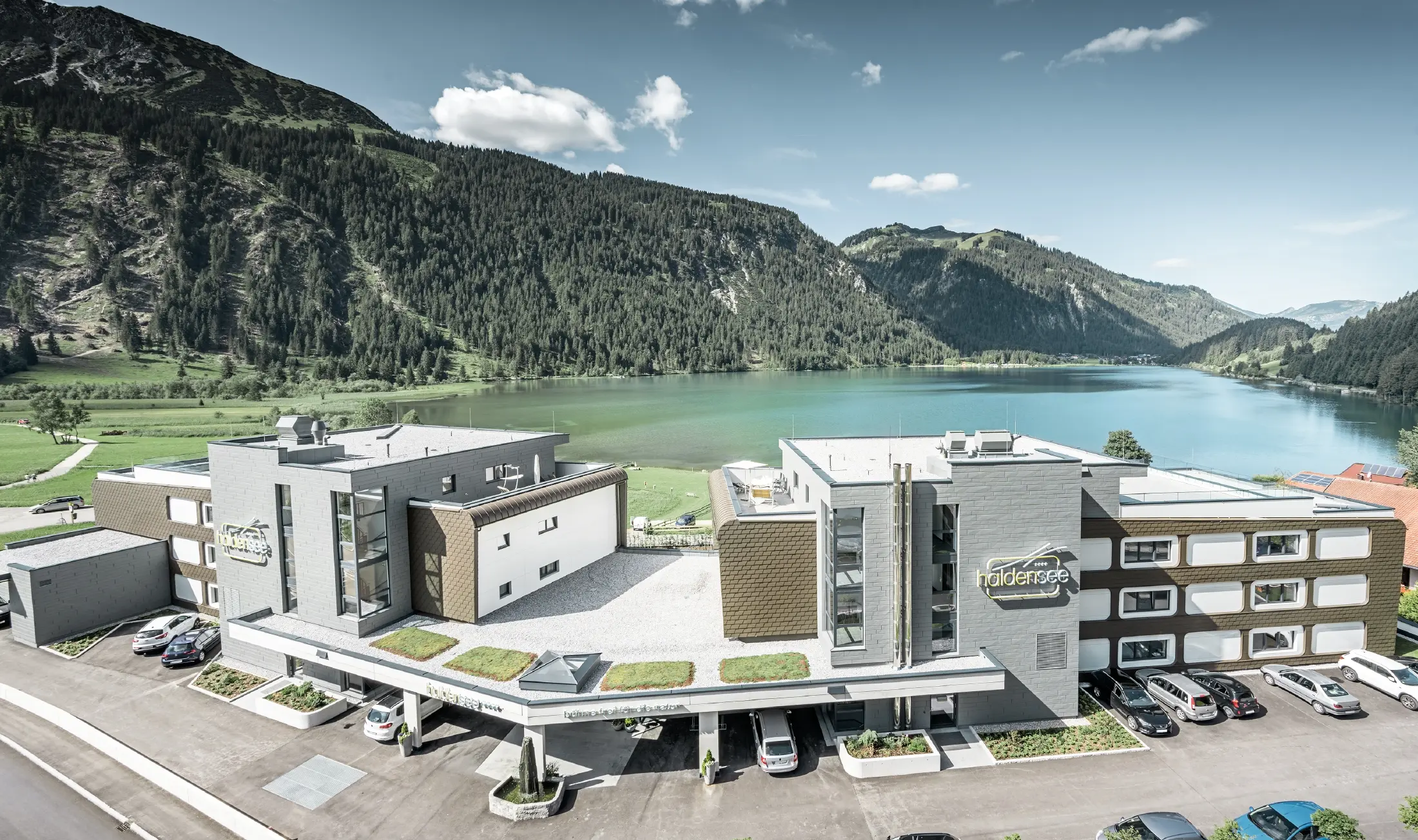 Hotel am Haldensee mit  Aluminium Verbundplatten, Wandschindeln, Prefalz und Siding.X, hinter dem Hotel sind Berge und ein See zu sehen