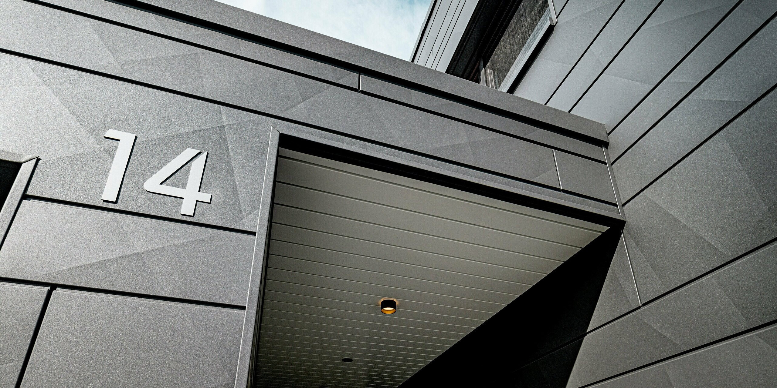 Detailansicht des überdachten Eingangs eines modernen Einfamilienhauses in Heidenheim mit PREFA Siding.X Fassade in P.10 Dunkelgrau. Die unterschiedlich gekanteten Aluminiumpaneele erzeugen ein markantes geometrisches Muster, das im Licht ein lebendiges Spiel aus Schatten und Reflexionen bildet. Die klar ausgeführte Hausnummer „14“ hebt sich präzise von der robusten Oberfläche ab. Die schützende Überdachung mit eingelassenen Spots unterstreicht die hochwertige Verarbeitung und zeigt die langlebige, witterungsbeständige Qualität der PREFA Aluminiumlösung.
