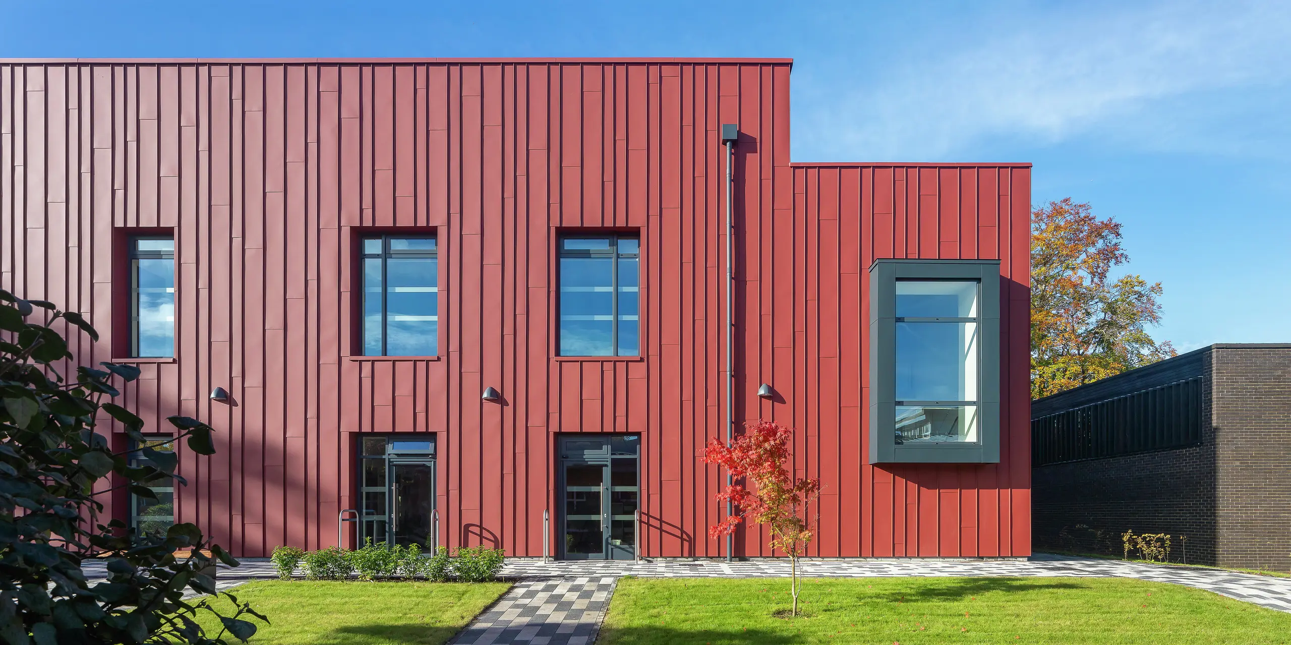 Moderne Architektur mit PREFALZ: Die Fassade des Soldiers' Centre in Pirbright beeindruckt mit einer eleganten Stehfalzverkleidung aus PREFALZ in P.10 Oxydrot. Die stilvolle Fensterrahmung in P.10 Anthrazit setzt gezielt Akzente und verleiht dem Gebäude ein dynamisches Erscheinungsbild. Langlebig, ästhetisch und nachhaltig - PREFA Aluminiumprodukte für innovative Baukonzepte.