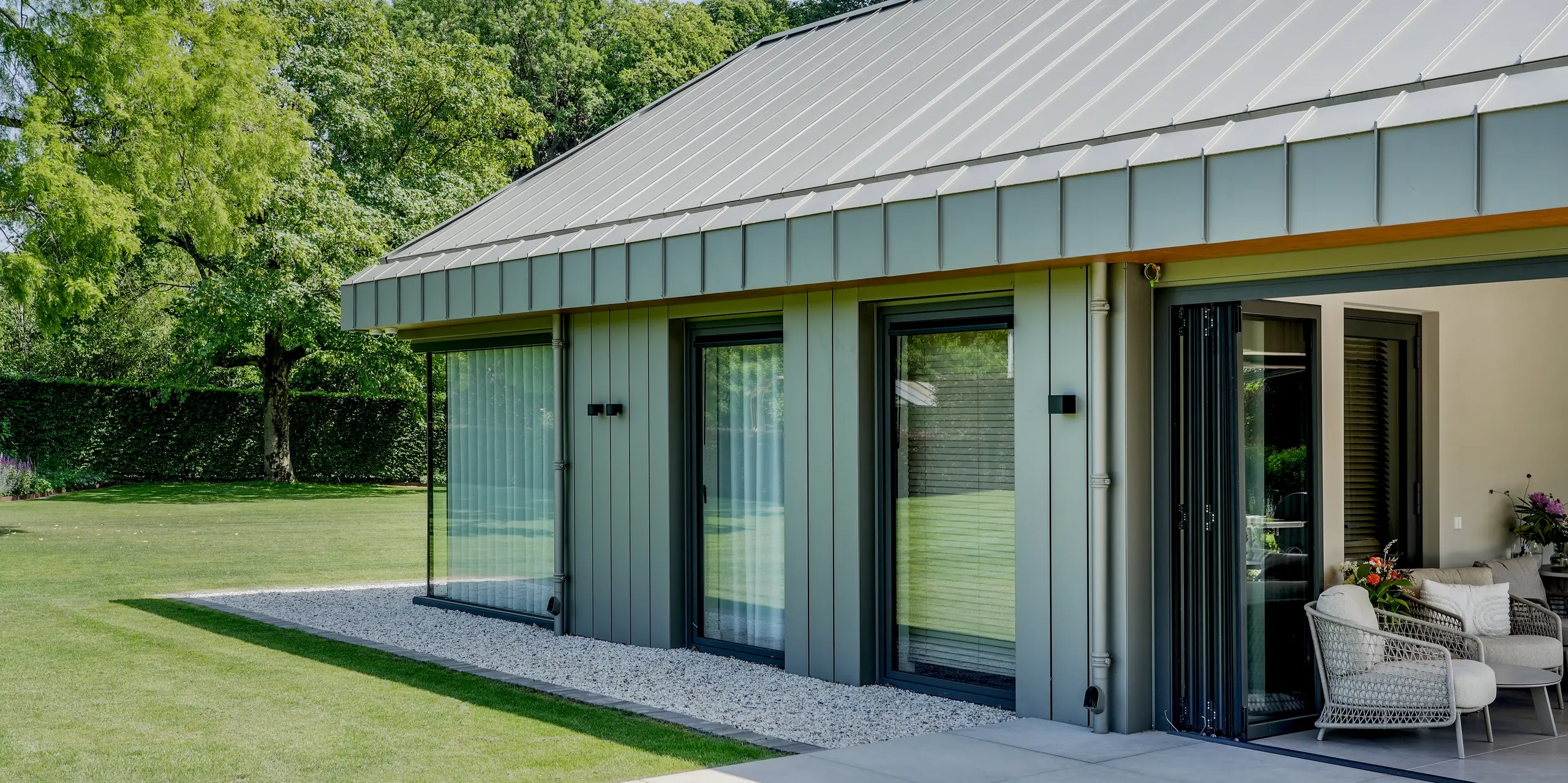 Moderner Anbau eines Einfamilienhauses in Putten mit PREFALZ Aluminiumdach und -fassade, eingebettet in eine üppige Gartenlandschaft. Die Winkelstehfalze, die großflächigen Verglasungen und die elegante Metallhülle schaffen einen eindrucksvollen Blickfang. PREFA Aluminium überzeugt durch langlebige Qualität, hohe Witterungsbeständigkeit und die harmonische Verbindung von Architektur und Natur.