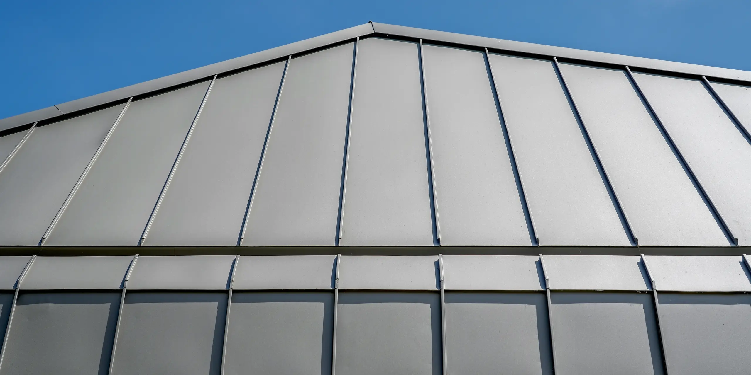Detailaufnahme des modernen Erweiterungsbaus in Putten: Die markante PREFALZ Aluminiumverkleidung mit präzisen Winkelstehfalzen zieht den Blick zur klar geformten Dachkante vor tiefblauem Himmel. Dach und Fassade verschmelzen zu einer eleganten Einheit. Das langlebige, wartungsarme PREFA Aluminium überzeugt mit hochwertiger Optik, Witterungsbeständigkeit und zeitloser Architektur.