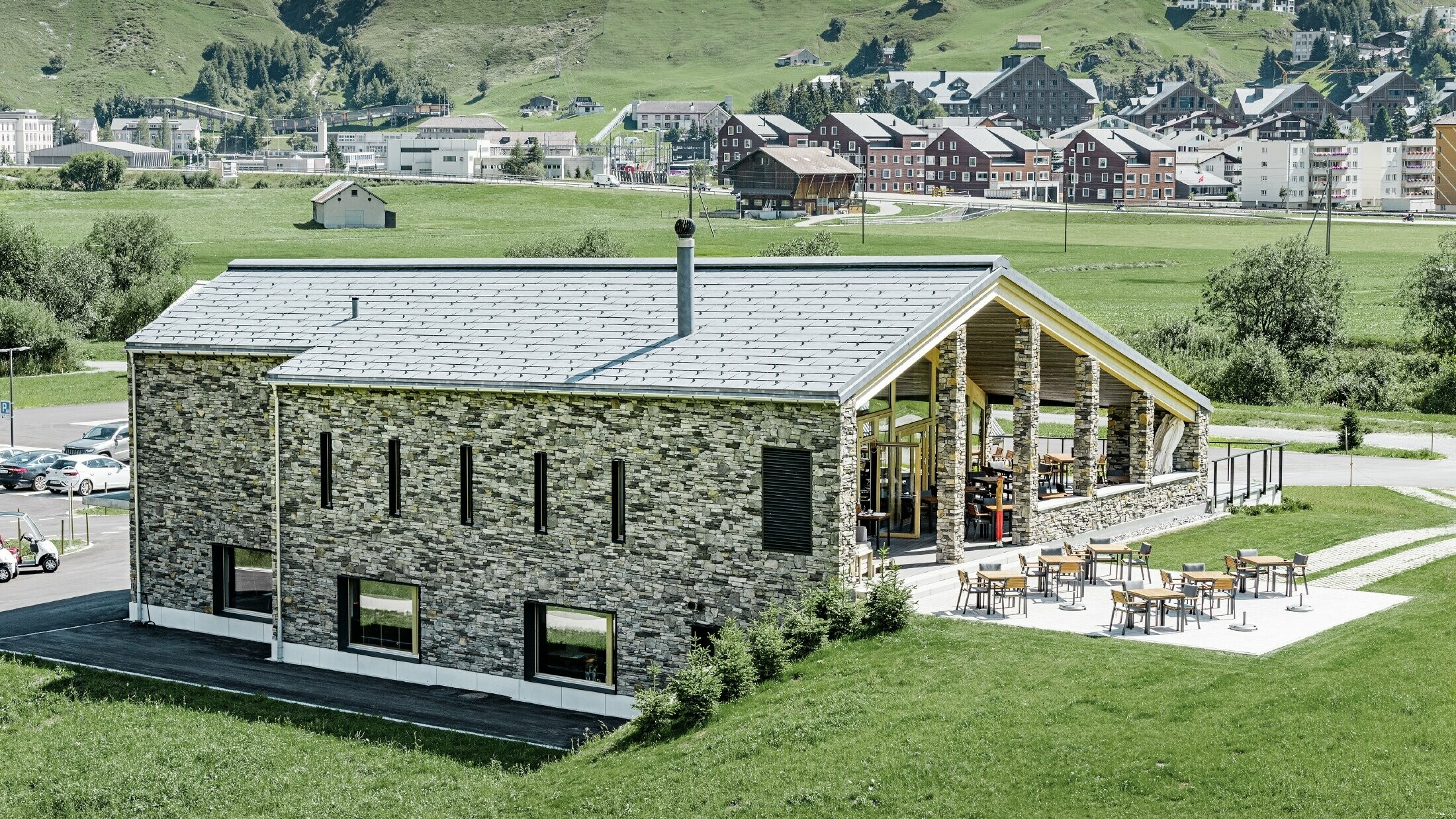 Il moderno punto di ritrovo del campo da golf a Andermatt con una facciata in pietra e il pannello per coperture FX.12 PREFA in alluminio in grigio pietra