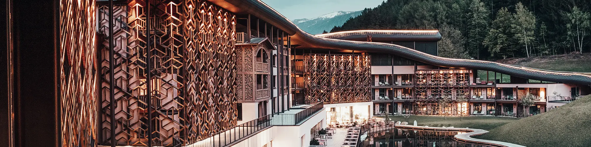 Abendaufnahme, warmes Licht beleuchtet die Holzfassade und das Aluminiumdach, unten die Terrasse mit Sitzgelegenheiten, davor der See, im Hintergrund der dunkelgrüne Wald