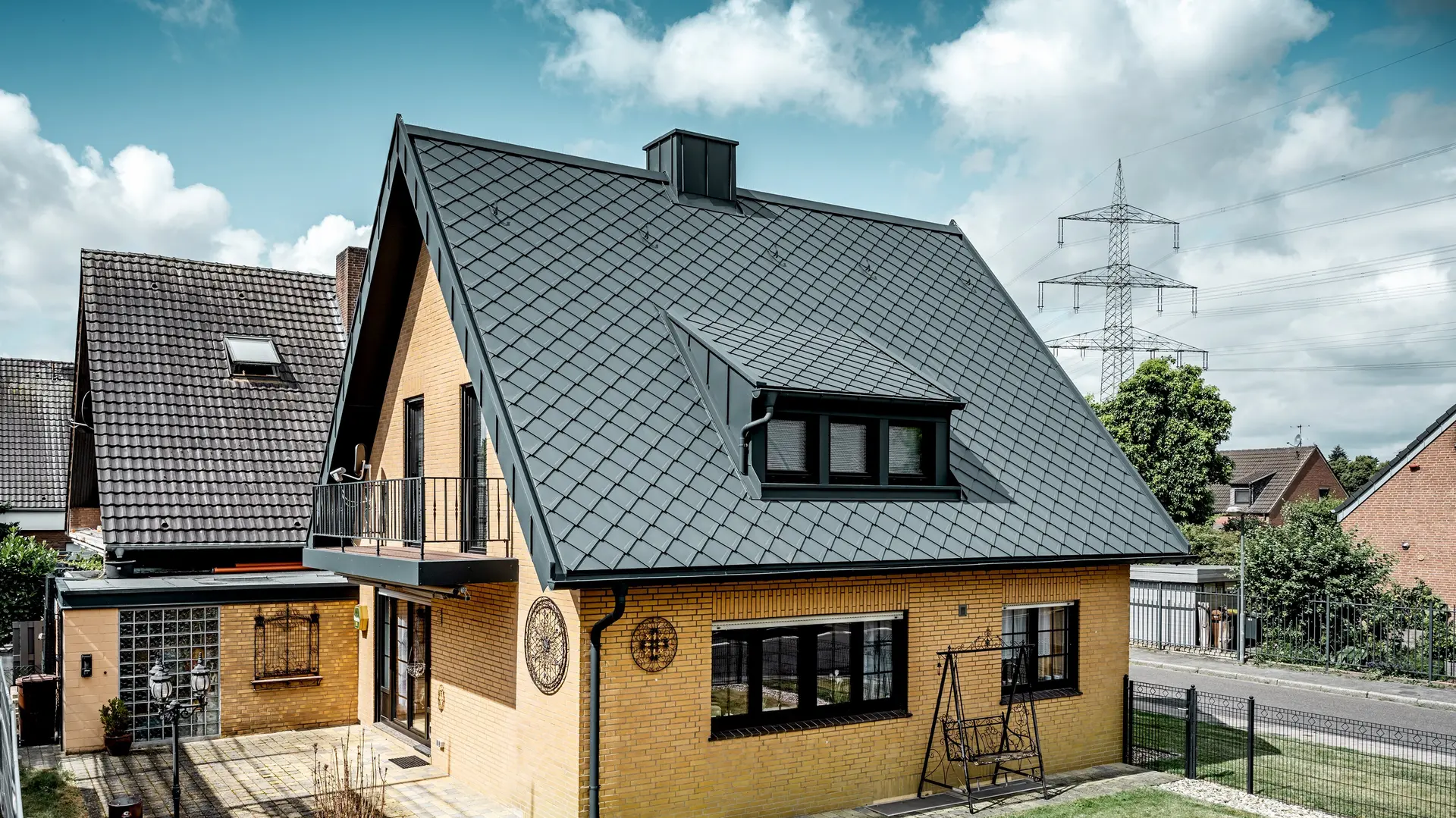 Casa unifamiliare in mattoni gialli dopo la ristrutturazione del tetto con PREFA scaglia 29 × 29 in grigio scuro; tetto a due falde ripido con copertura nuova, motivo a rombi, abbaino e camino integrati, terrazza e giardino in primo piano.