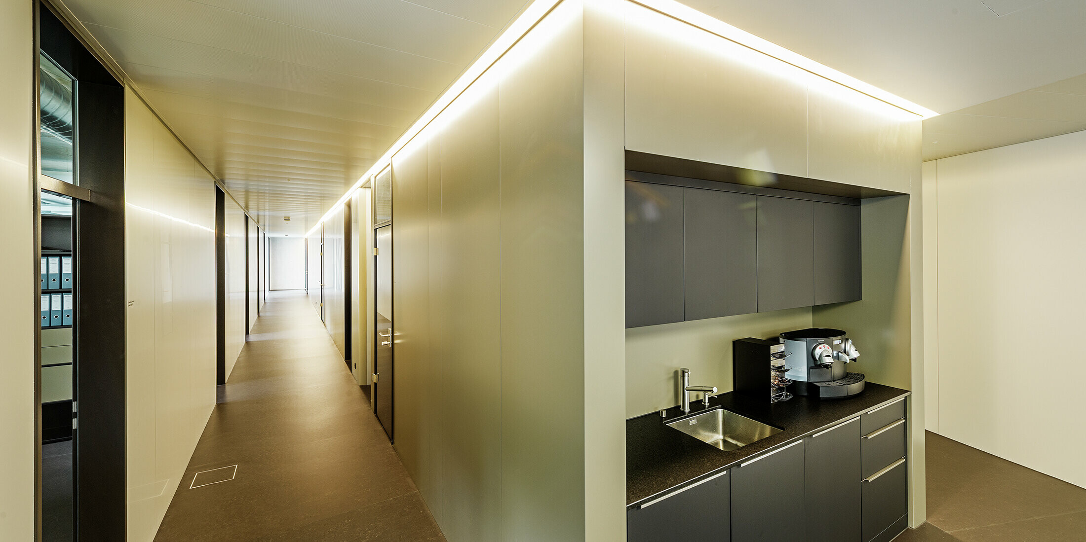 In den modernen Büroräumlichkeiten der Firma Guggisberg Dachtechnik in Wabern sorgt die integrierte Teeküche für Funktionalität und Design. Die Wandverkleidungen nehmen die edle Bronzeoptik der PREFA Aluminiumfassade auf und fügen sich nahtlos in das architektonische Konzept ein. Die Innenraumgestaltung mit indirekter Beleuchtung schafft ein angenehmes Raumgefühl.