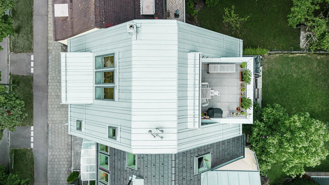 Vista aerea di una casa a schiera ristrutturata in chiave moderna. La casa ha il tetto e la facciata del piano superiore in alluminio Prefalz silver metallizzato, che enfatizza le linee orizzontali. Una terrazza sul tetto con arredi da esterno e piante crea un'oasi verde urbana. La facciata del seminterrato è di un colore più scuro, che crea un piacevole contrasto. La casa è circondata da un'area verde ben curata e da sentieri pavimentati che sottolineano la chiara struttura dell'edificio.