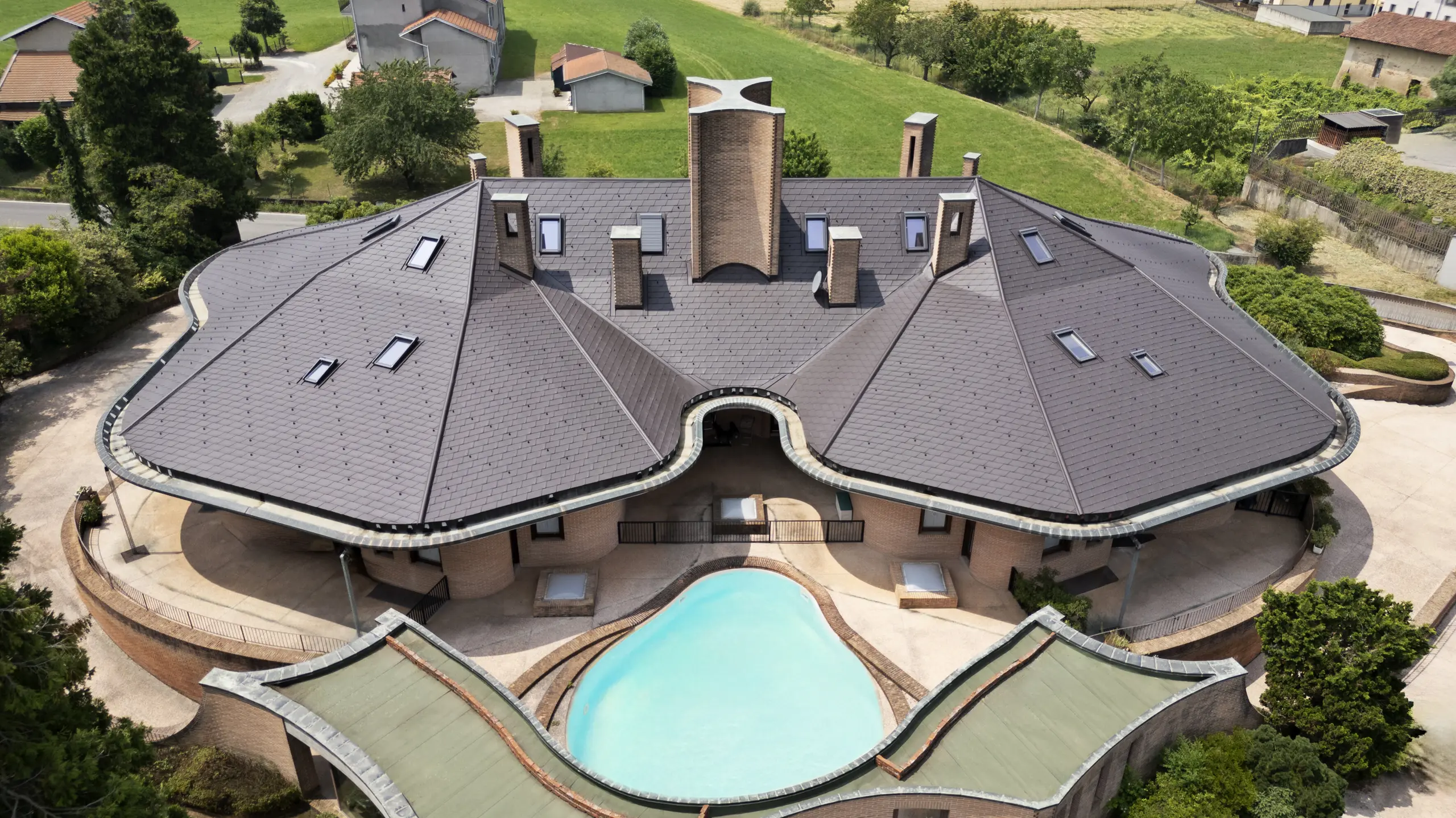 Foto dall'alto della ristrutturazione di una villa degli anni ’90 a Oleggio, in provincia di Novara. Il caratteristico tetto a forma di farfalla è stato rivestito con scandole PREFA DS.19 in alluminio, nel colore P.10 testa di moro, valorizzando la particolare geometria della copertura e l’architettura dell’edificio. Perfettamente integrato nel paesaggio rurale attorno e con una bellissima piscina.