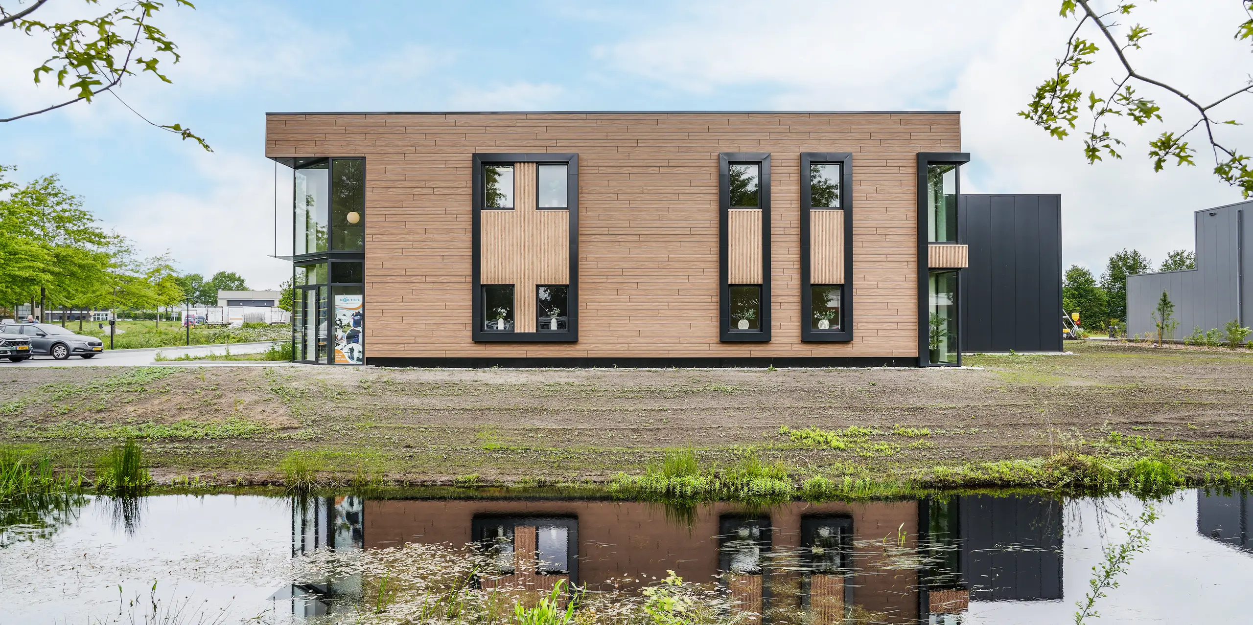 Das moderne Firmengebäude der Dokter Schoonmaakorganisatie in Wolvega spiegelt sich eindrucksvoll im ruhigen Flus davor. Die walnussbraune PREFA Fassade mit Sidings in authentischer Holzoptik prägt den kubischen Baukörper und bildet einen attraktiven Kontrast zu den dunklen Fensterrahmen. Großzügige Verglasungen, klare Linien und die naturnahe Umgebung unterstreichen die hochwertige, zeitgemäße Architektur.