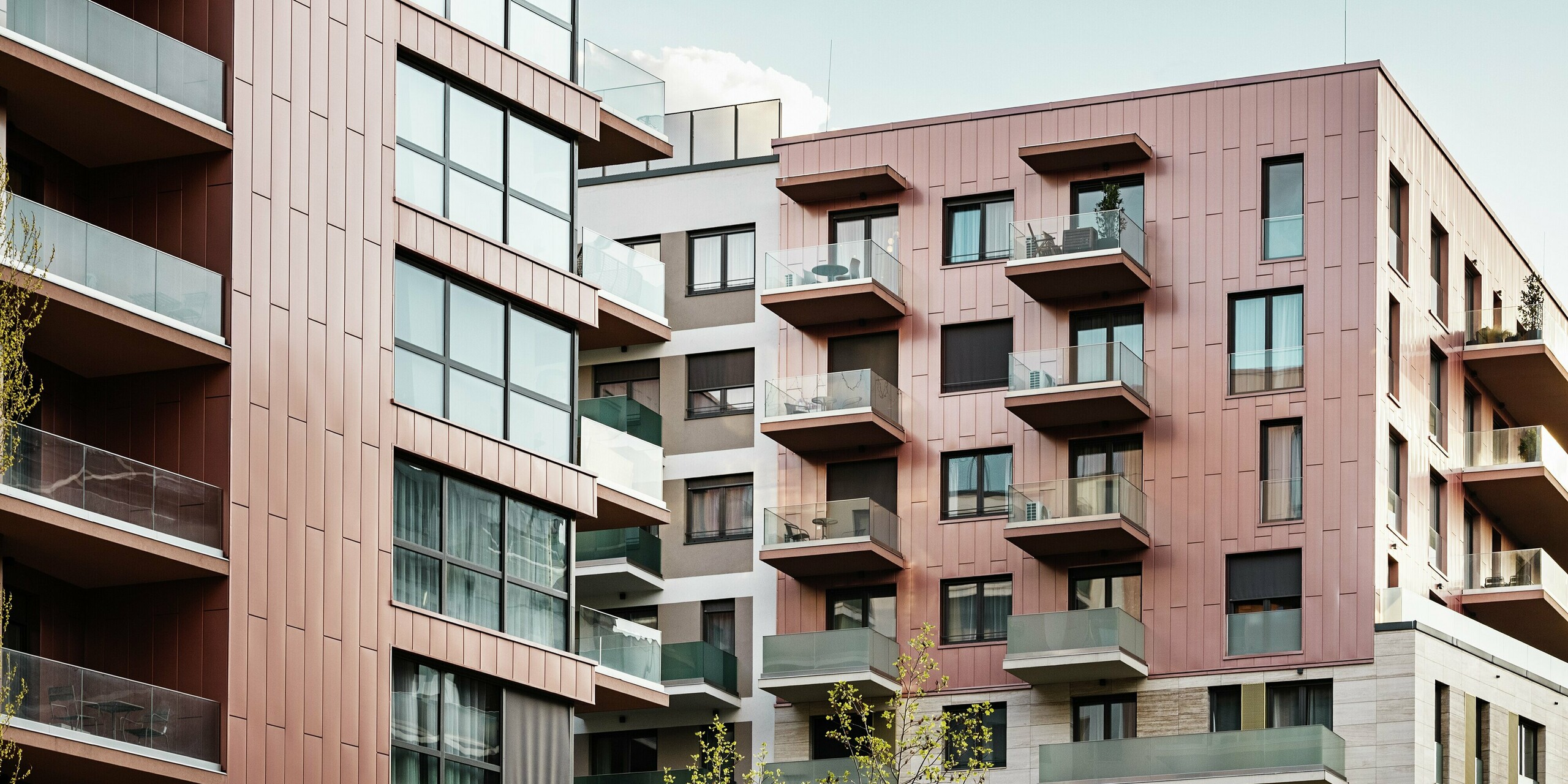 Fassadendetail des Wohnkomplexes Grand Corvin im revitalisierten Corvin-Viertel von Budapest. Die vertikal verlegten PREFA Siding Fassadenpaneele in der Sonderfarbe Lachsmetallic prägen das moderne Erscheinungsbild der oberen Gebäudeetagen. In Kombination mit großflächigen Glasfronten und Naturstein schafft die langlebige Aluminiumverkleidung ein harmonisches und hochwertiges Stadtbild.