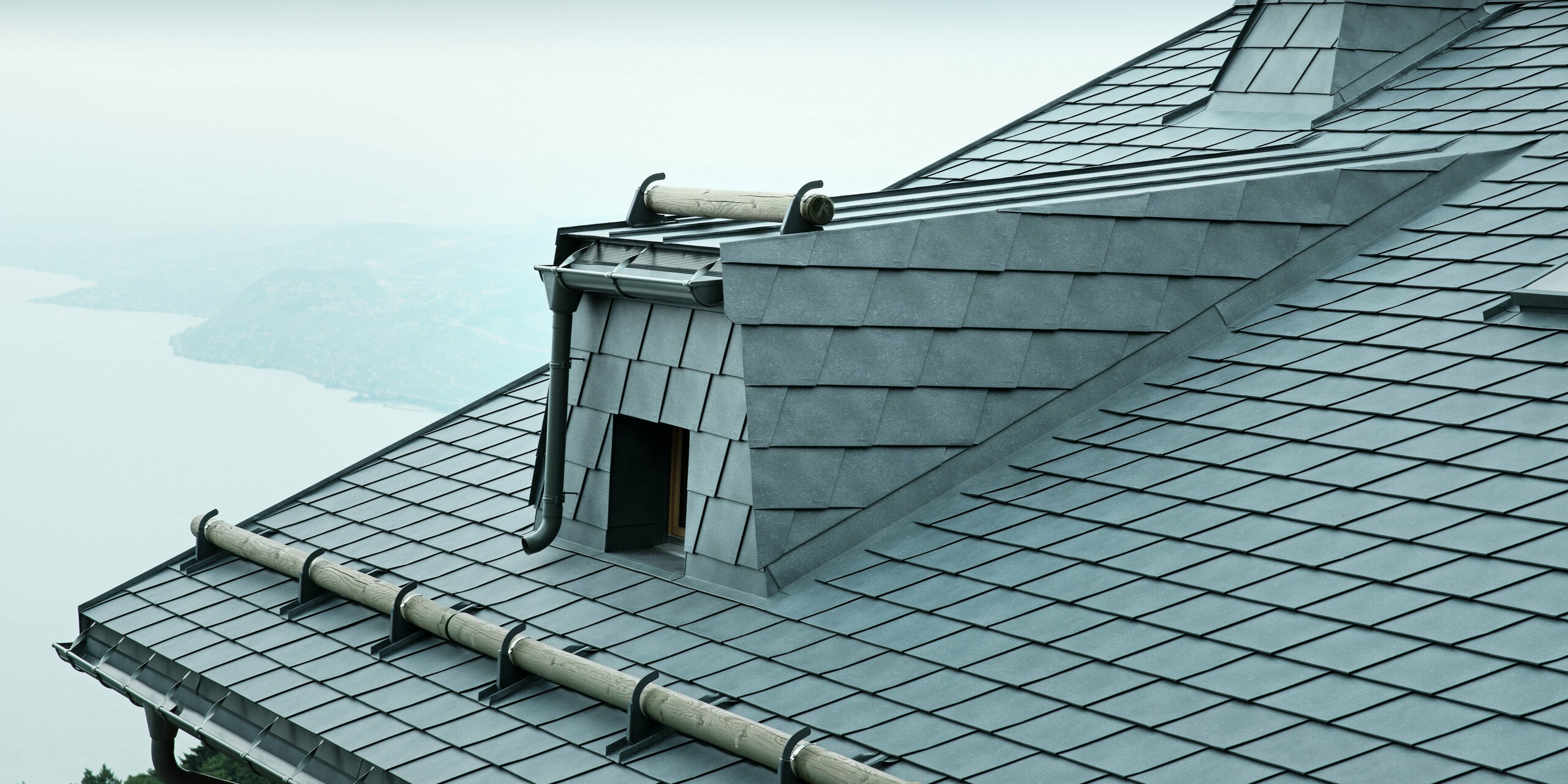 Die Dachansicht dieser modernen Holz-Lodge in der Schweiz zeigt steingraue PREFA Aluminium-Dachschindeln und eine große Dachgaube. Das langlebige Aluminiumdach schützt das Gebäude vor den extremen Wetterbedingungen am Genfersee. Der raffinierte Schneefang aus Rundholz und der markante Kamin betonen das einzigartige Design der Architektur.