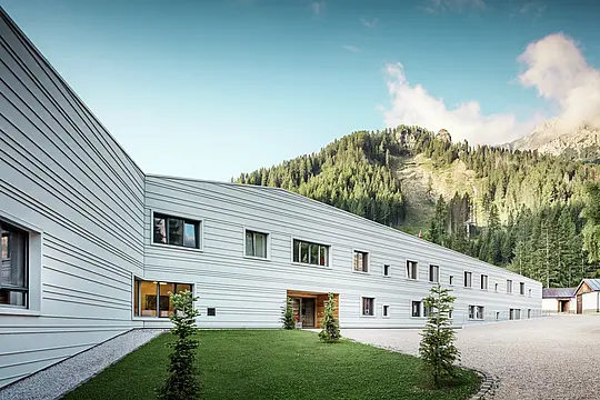 L'alluminio incontra la precisione: un dialogo armonioso tra tecnologia e natura. Davanti all'impressionante scenario delle Dolomiti, il “Villaggio Alpino Tempesti” si presenta con linee pulite e un design moderno. La facciata in PREFALZ nel colore P.10 bianco PREFA non è solo estremamente resistente, ma grazie al suo colore chiaro garantisce anche un microclima piacevole, poiché riflette i raggi del sole. Le aree verdi e gli elementi in legno nella zona d'ingresso dell'edificio militare creano un'atmosfera sorprendentemente accogliente.
