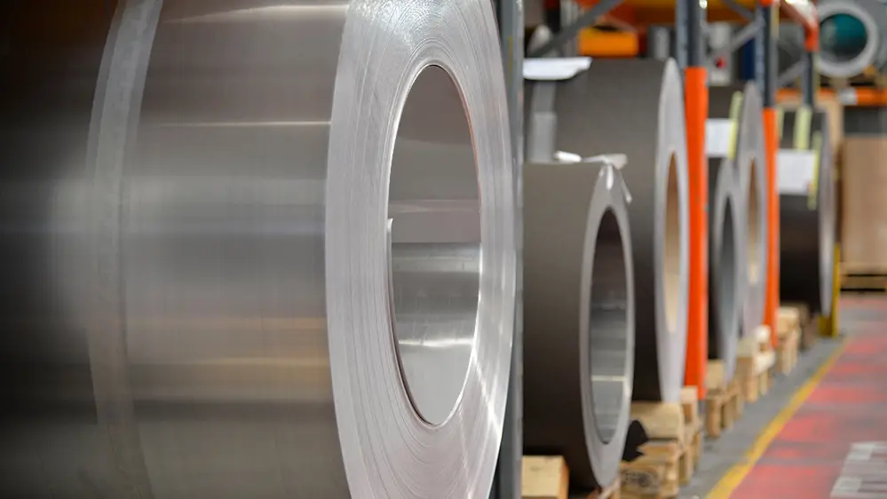 Bobine di alluminio (coil) stoccate su pallet di legno; in primo piano è visibile un coil di alluminio naturale. Dietro, altri nastri e fogli di alluminio.