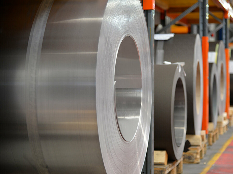 Bobine di alluminio (coil) stoccate su pallet di legno; in primo piano è visibile un coil di alluminio naturale. Dietro, altri nastri e fogli di alluminio.