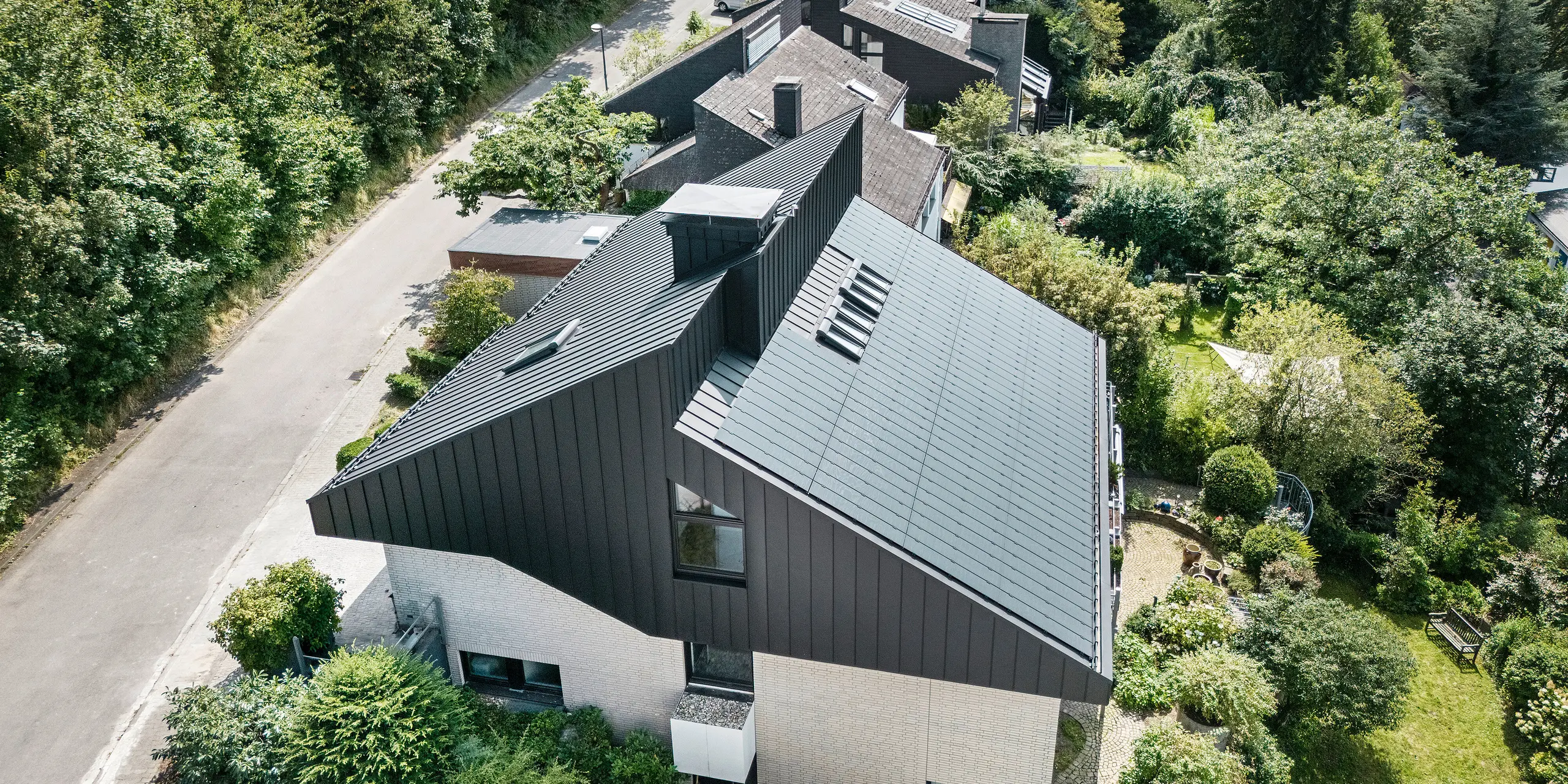 Schrägansicht eines modernen Einfamilienhauses in Meschede, Deutschland. Das Satteldach ist mit PREFALZ P.10 Schwarz gedeckt. Das Aluminiumdach setzt einen optischen Akzent und bietet dem Gebäude langfristigen Schutz. Die präzise Doppeldachkonstruktion erstreckt sich vom Dach über die Fassade bis zur Vorderseite. Umgeben von üppigem Grün und einer ruhigen Straße fügt sich das energieeffiziente Haus mit innovativen PREFALZ-Solarmodulen harmonisch in die natürliche Umgebung ein.