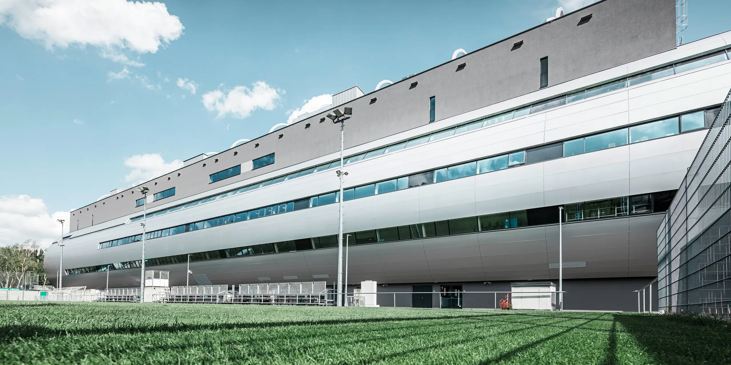 Das Allianz Stadion von SK Rapid Wien, abgebildet vom Trainingsplatz, zeigt die glänzende Fassade aus PREFA Aluminium Verbundplatten in Silbermetallic. Die harmonische Verschmelzung von funktionaler Sportstätte und eleganter Architektur wird durch die Fensterreihen betont. Die grüne Wiese des Trainingsplatzes am unteren Bildrand wird durch die Betreuerbänke und den Zaun abgegrenzt. Am Zaus sind zudem hohe Flutlichtmasten zu erkennen, die eine Ausübung des Trainings zu jeder Tageszeit ermöglichen. Der blaue Himmel komplementiert das professionelle Foto.