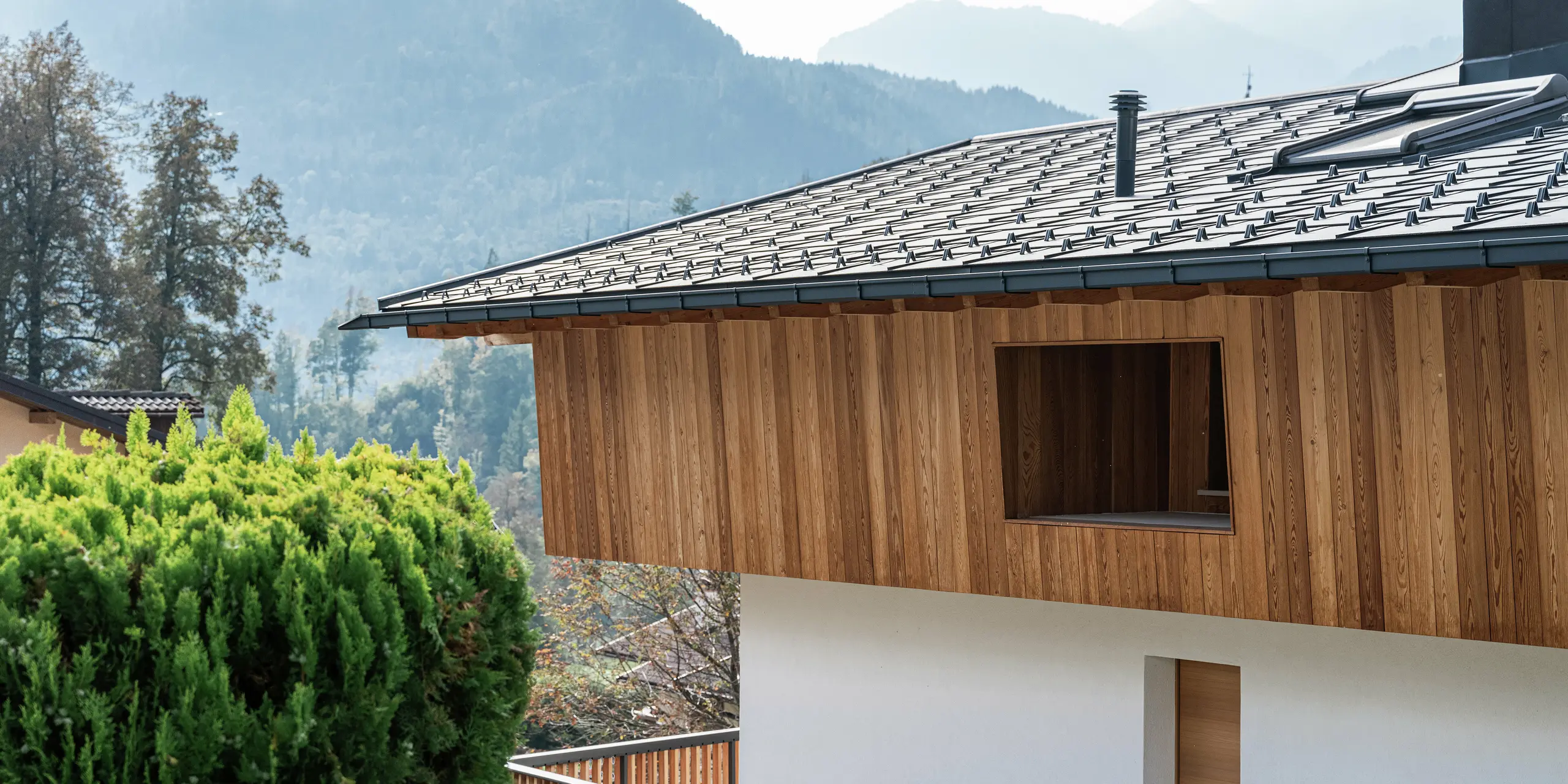 Detailansicht eines traditionellen Einfamilienhauses in Agordo, Italien, mit einer natürlichen Holzfassade und robusten PREFA Dachplatten R.16 in P.10 Anthrazit. Das widerstandsfähige Aluminiumdach mit Schneestoppern sorgt für optimalen Witterungsschutz. Die Kombination aus naturbelassener Holzfassade im Obergeschoss und weißer Putzfassade im Erdgeschoss verleiht dem Haus ein warmes, modernes Erscheinungsbild. Die klare Linienführung und die hochwertigen Materialien fügen sich harmonisch in die alpine Umgebung ein.