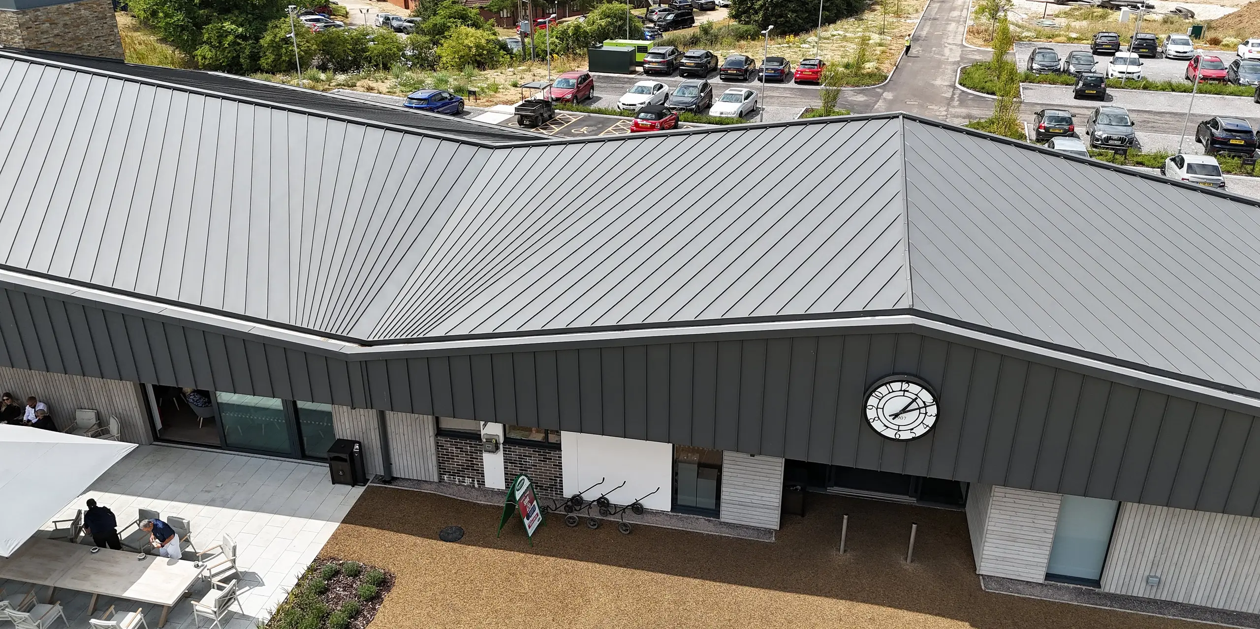 Die beeindruckende Dachkonstruktion des Basingstoke Golf Clubs in Hampshire, mit PREFALZ Stehfalzbahnen in P.10 Dunkelgrau, zieht sofort die Aufmerksamkeit auf sich. Die geschwungene Form des Daches und die präzise Anordnung der Stehfalzbahnen schaffen ein markantes, modernes Design, das sich harmonisch in die Umgebung einfügt. Auf der unteren Seite der Fassade sind Holzpaneele zu sehen, die einen natürlichen Kontrast zur metallischen Oberfläche bilden. Der Außenbereich mit Sitzplätzen ergänzt das Bild eines modernen, einladenden Clubhauses.