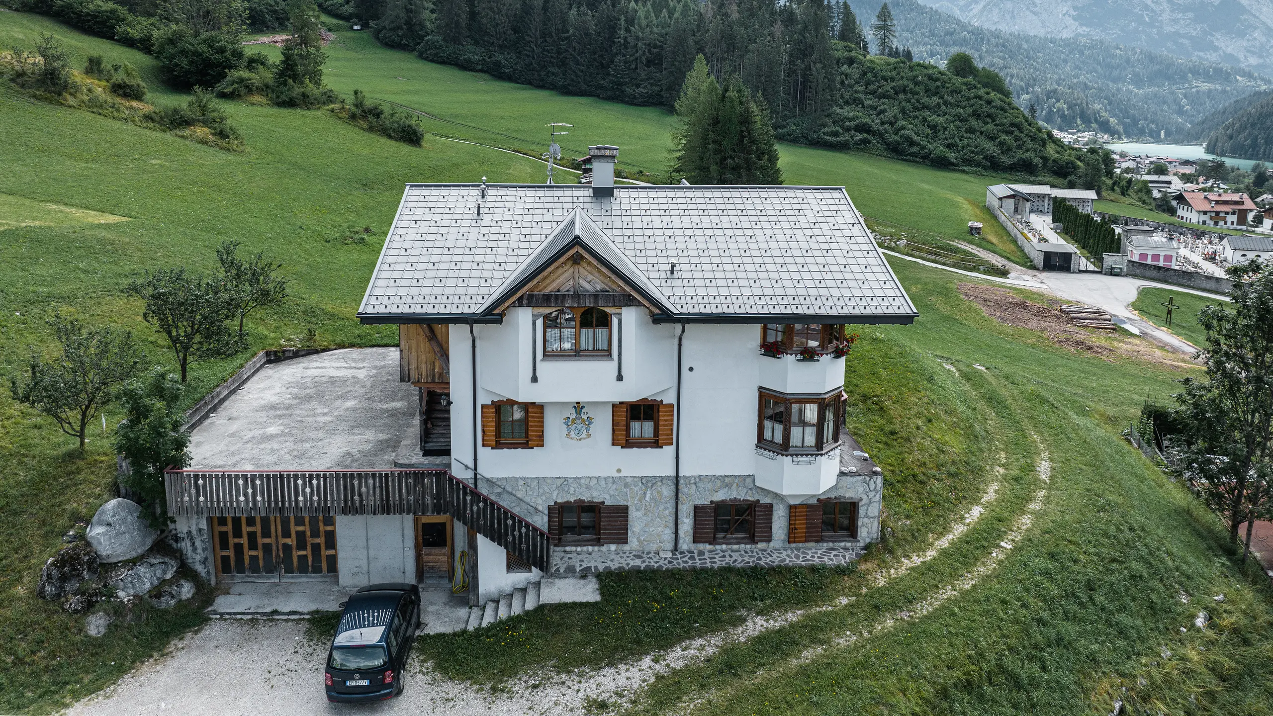Vista a volo d'uccello dell'ingresso del tradizionale rifugio alpino di Auronzo di Cadore dopo la ristrutturazione del tetto con tegole PREFA in alluminio P.10 grigio chiaro. L'edificio, con il suo caratteristico fascino alpino e il moderno tetto in lamiera, si inserisce armoniosamente nel paesaggio verde e nel suggestivo panorama montano sullo sfondo, dimostrando una riuscita combinazione di funzionalità e architettura regionale.