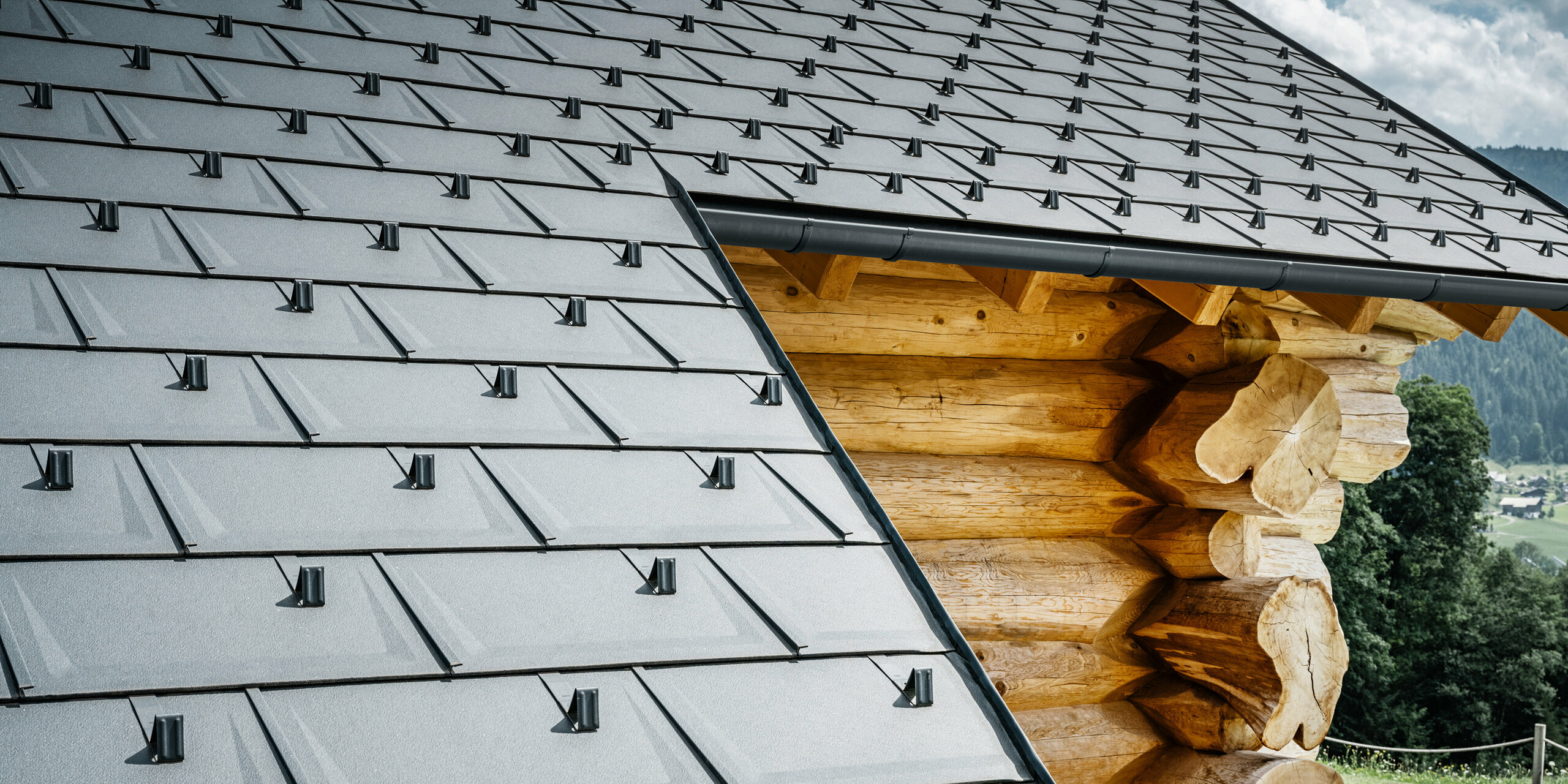 Detailaufnahme eines Daches mit PREFA Dachplatten R.16 in der Farbe P.10 Anthrazit auf einem Blockhaus in Gosau, Oberösterreich. Die präzise Verarbeitung und Montage der robusten Dachplatten zeigt die hohe Qualität der PREFA Aluminiumprodukte sowie die perfekte Integration moderner Bauelemente aus Aluminium in traditionelle Gebäudetypen aus Österreich. Im Vordergrund sind die charakteristischen Schneestopper zu sehen, die Sicherheit im alpinen Winter gewährleisten. Dieses Bild fängt das harmonische Zusammenspiel von innovativen PREFA Dachsystemen mit der natürlichen Ästhetik des Holzblockhauses ein.