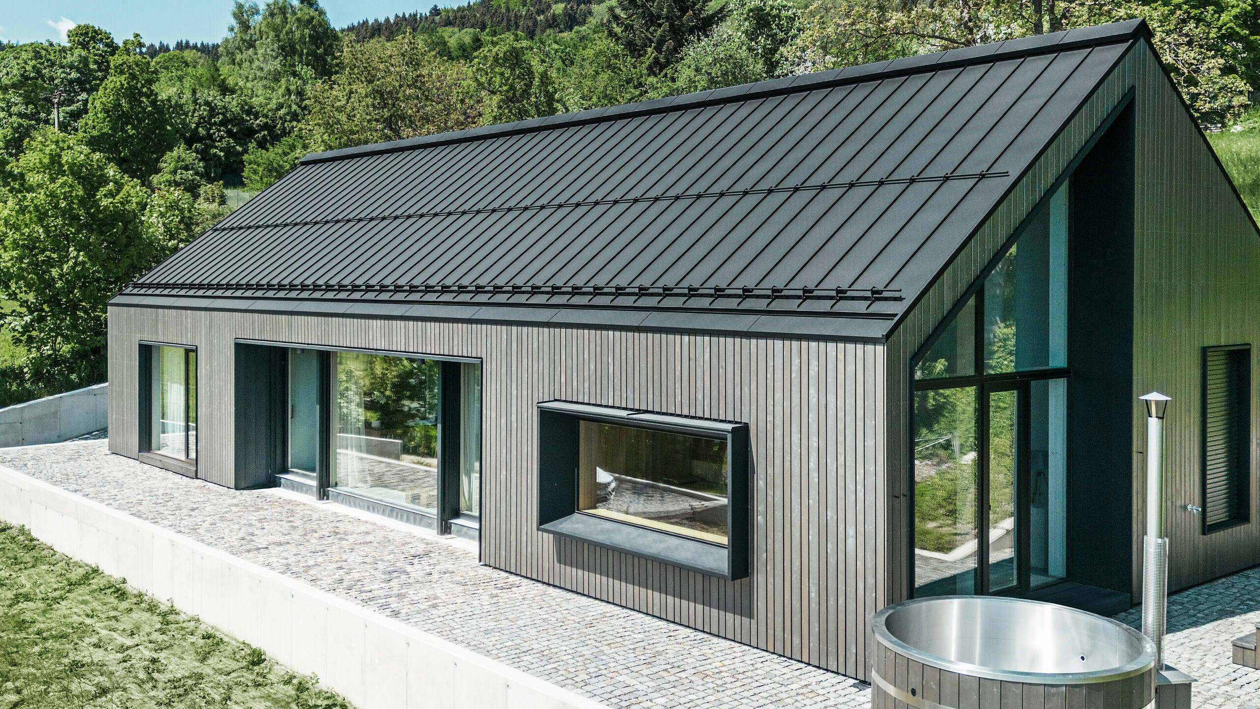 Ein modernes Holzmassivhaus in Kuncice (CZ) wurde mit großen Fensterflächen ausgestattet. Das dunkle Holzmassivhaus verfügt über ein Dachsystem aus PREFALZ in der Farbe P. 10 Schwarz. Im hinteren Außenbereich steht ein Whirlpool mit einer Holzverkleidung.