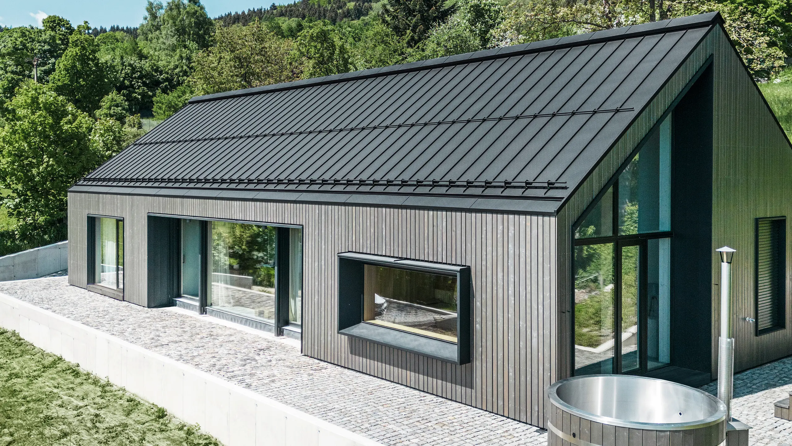 Ein modernes Holzmassivhaus in Kuncice (CZ) wurde mit großen Fensterflächen ausgestattet. Das dunkle Holzmassivhaus verfügt über ein Dachsystem aus PREFALZ in der Farbe P. 10 Schwarz. Im hinteren Außenbereich steht ein Whirlpool mit einer Holzverkleidung.