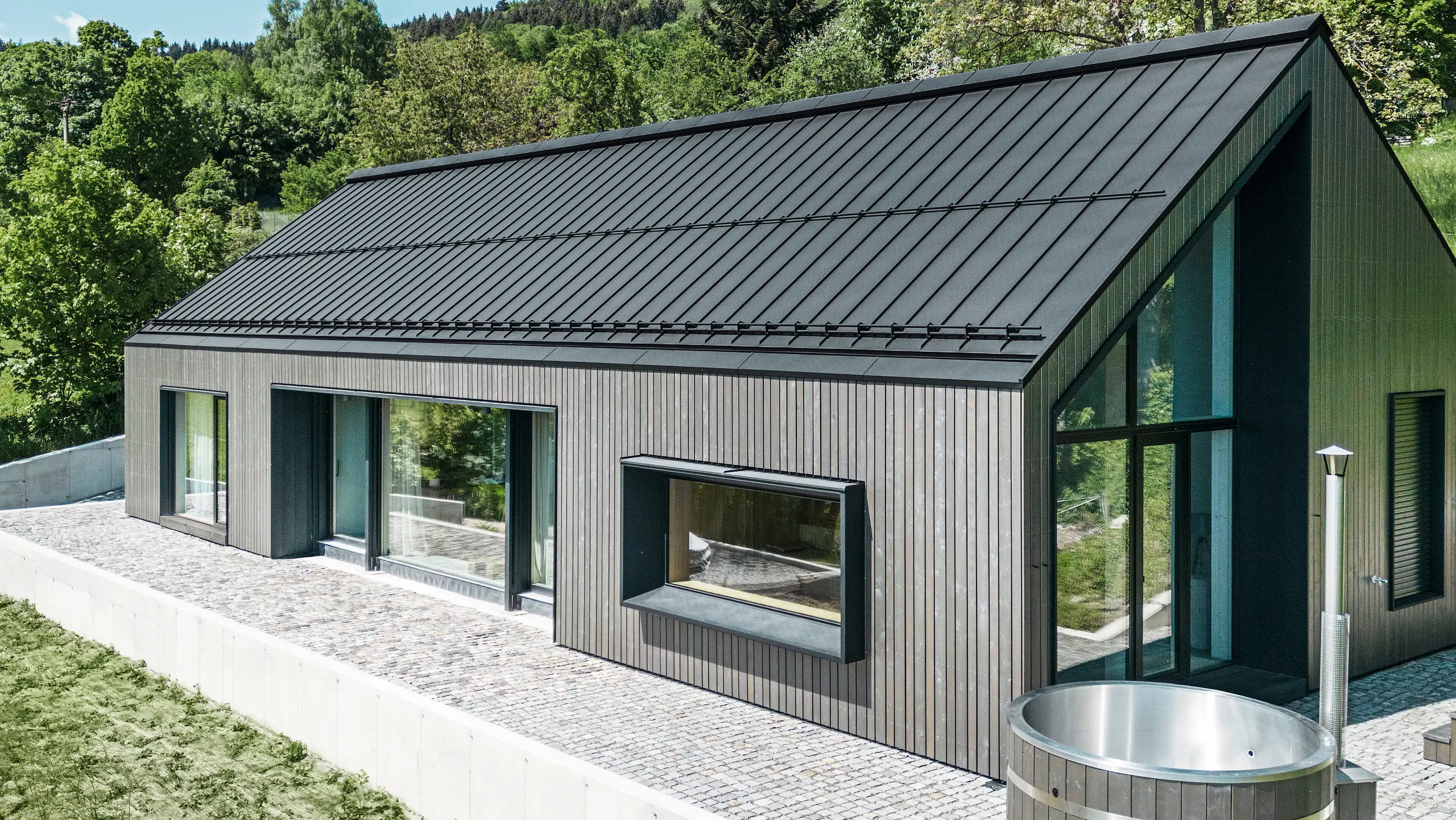 Ein modernes Holzmassivhaus in Kuncice (CZ) wurde mit großen Fensterflächen ausgestattet. Das dunkle Holzmassivhaus verfügt über ein Dachsystem aus PREFALZ in der Farbe P. 10 Schwarz. Im hinteren Außenbereich steht ein Whirlpool mit einer Holzverkleidung.