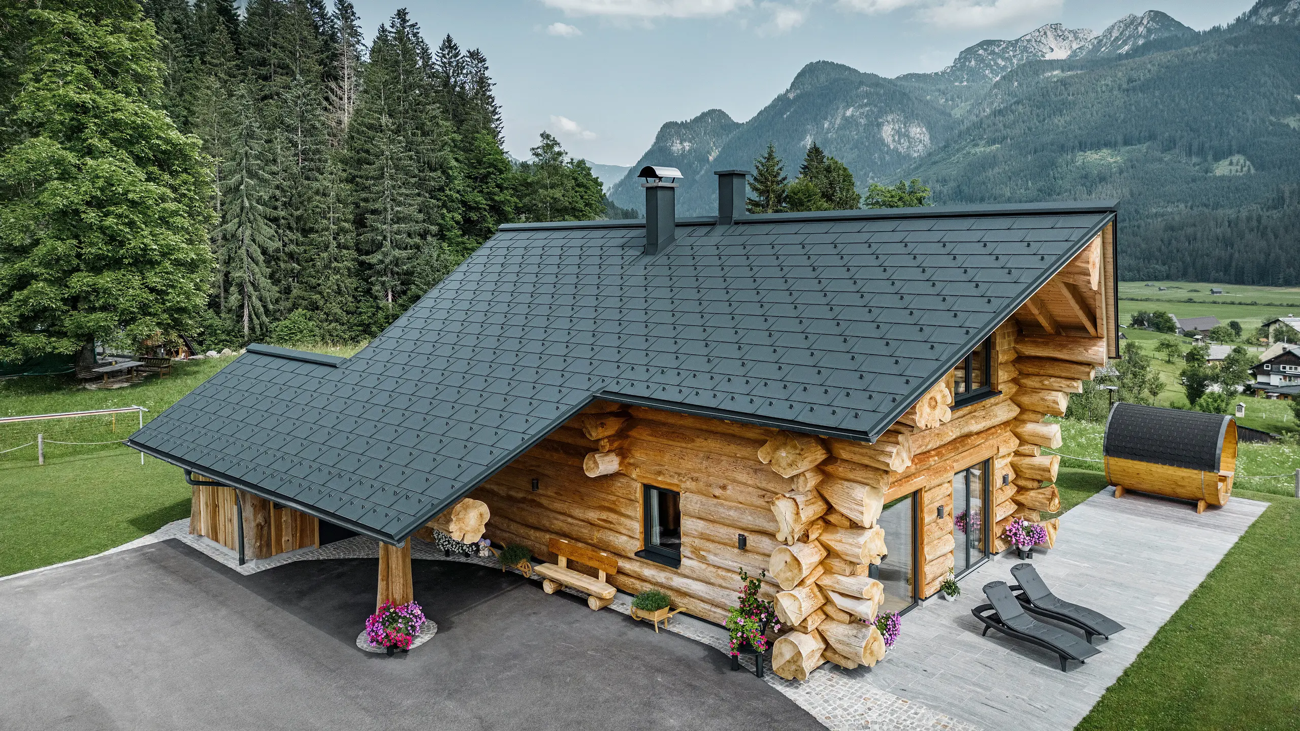 Schräge Ansicht eines malerischen Blockhauses in Gosau, Österreich, ausgestattet mit einem PREFA Dach aus anthrazitfarbenen Dachplatten R.16. Die Architektur kombiniert rustikales Blockhausflair mit modernen Elementen, umgeben von dichten grünen Wäldern und einer beeindruckenden Bergkulisse. Das Haus strahlt mit seiner ländlichen Eleganz und den präzise verlegten Dachelementen aus Aluminium, die für ihre Langlebigkeit und Witterungsbeständigkeit bekannt sind, eine einladende Atmosphäre aus.