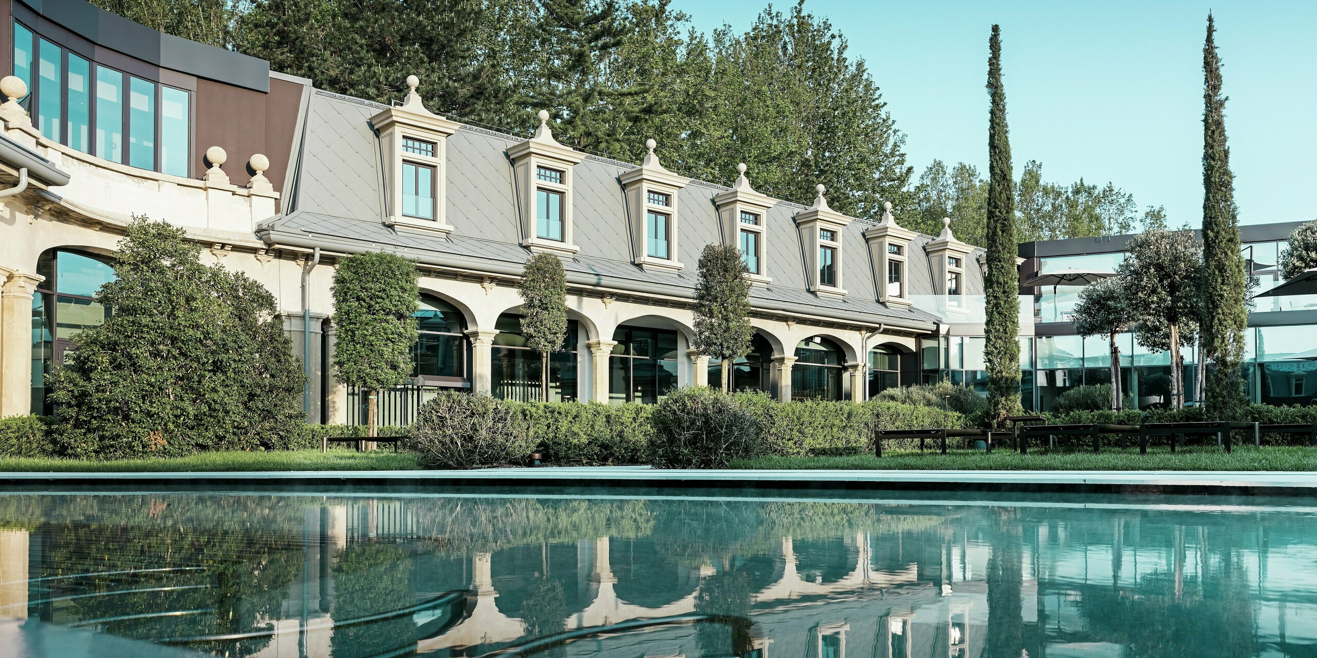 Blick über die ruhige Wasserfläche eines Pools auf die historische Fassade der Therme „De Montel – Terme Milano“. Die markante Dachgestaltung mit PREFA Dachrauten 44 × 44 in P.10 Hellgrau und den fein ausgearbeiteten Gauben spiegelt sich eindrucksvoll im Wasser. Umgeben von gepflegten Grünflächen, Zypressen und modernen Glasbauten entsteht ein harmonisches Zusammenspiel aus Tradition, Natur und zeitgemäßer Architektur.