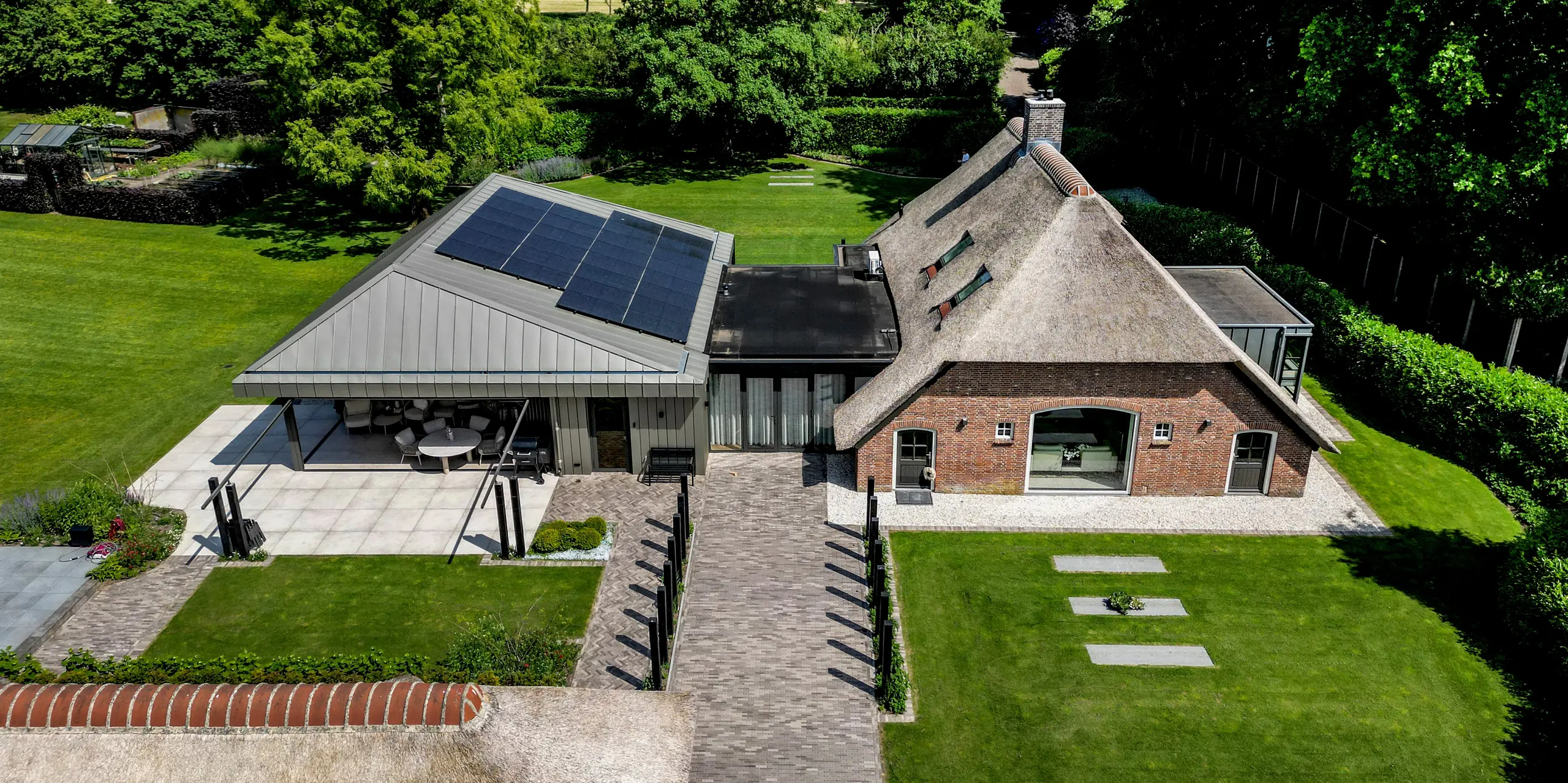 Spektakuläre Luftaufnahme des Einfamilienhauses in Putten, die den spannenden Kontrast zwischen traditionellem Reetdach und modernem PREFALZ Anbau zeigt. Die elegante Aluminiumhülle in P.10 Prefabronze, integrierte Solarmodule und großzügige Terrassenflächen setzen starke Akzente. PREFA Aluminium überzeugt durch langlebige Qualität, Witterungsbeständigkeit und die harmonische Verbindung von Architektur, Nachhaltigkeit und Natur.