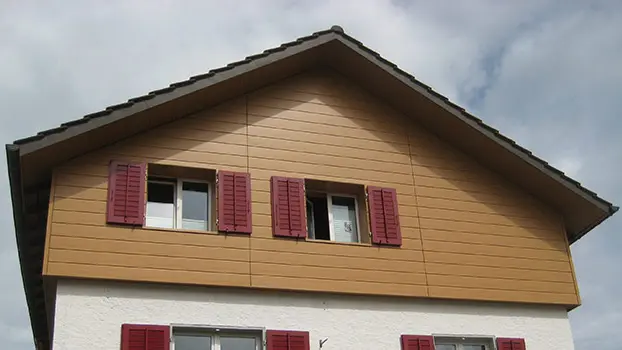 Rivestimento del timpano di una casa tradizionale con tetto a due falde. Il timpano è rivestito con doghe di rivestimento PREFA (rovere naturale) in posa orizzontale. Le finestre sono provviste di persiane rosse.