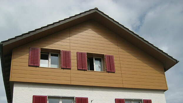 Rivestimento del timpano di una casa tradizionale con tetto a due falde. Il timpano è rivestito con doghe di rivestimento PREFA (rovere naturale) in posa orizzontale. Le finestre sono provviste di persiane rosse.