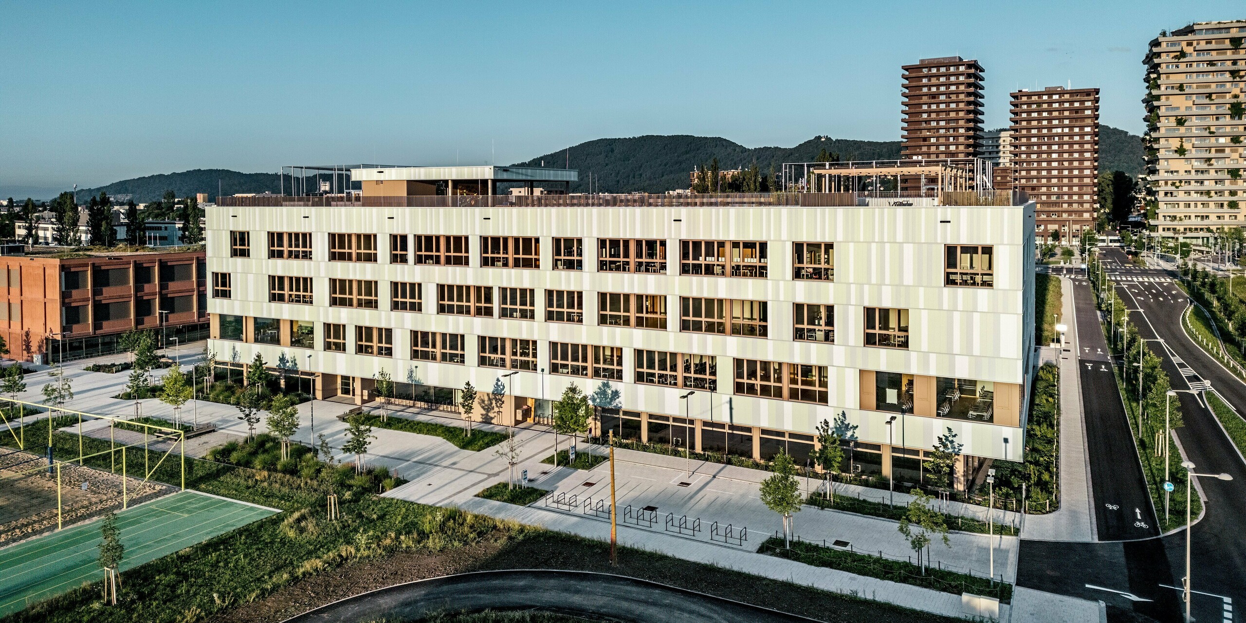 Das Bild zeigt die AHS Reininghaus in Graz aus der Vogelperspektive und hebt die moderne Architektur des Gebäudes hervor. Die mit PREFA Sidings in Sonderfarben verkleidete Fassade präsentiert sich in sanften Grüntönen und Weiß, was dem Gebäude eine frische, lebendige Ausstrahlung verleiht. Besonders auffällig sind die klar strukturierten, vertikal angeordneten Aluminiumprofile. Im Vordergrund erstreckt sich ein großzügiger Außenbereich mit grünen Rasenflächen, einem Sportplatz und Fahrradständern, der die Funktionalität des Campus unterstreicht. Die moderne Anordnung von Wegen und Pflanzen schafft ein angenehmes, einladendes Umfeld. Die Architektur und Materialwahl harmonieren perfekt mit der modernen Infrastruktur der Umgebung und machen die AHS Reininghaus zu einem markanten Beispiel für die Architektur zeitgenössischer Bildungseinrichtungen.