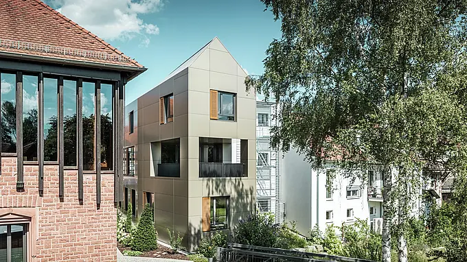 L'immagine mostra il moderno municipio di Alzenau, la cui facciata è realizzata con pannelli compositi in alluminio PREFA color bronzo. Il tetto a punta e le finestre dalla struttura chiara con persiane in legno conferiscono all'edificio un'estetica elegante e contemporanea. In primo piano si vede un'area verde con un sentiero lastricato e alberi, che completa armoniosamente il design urbano dell'edificio.