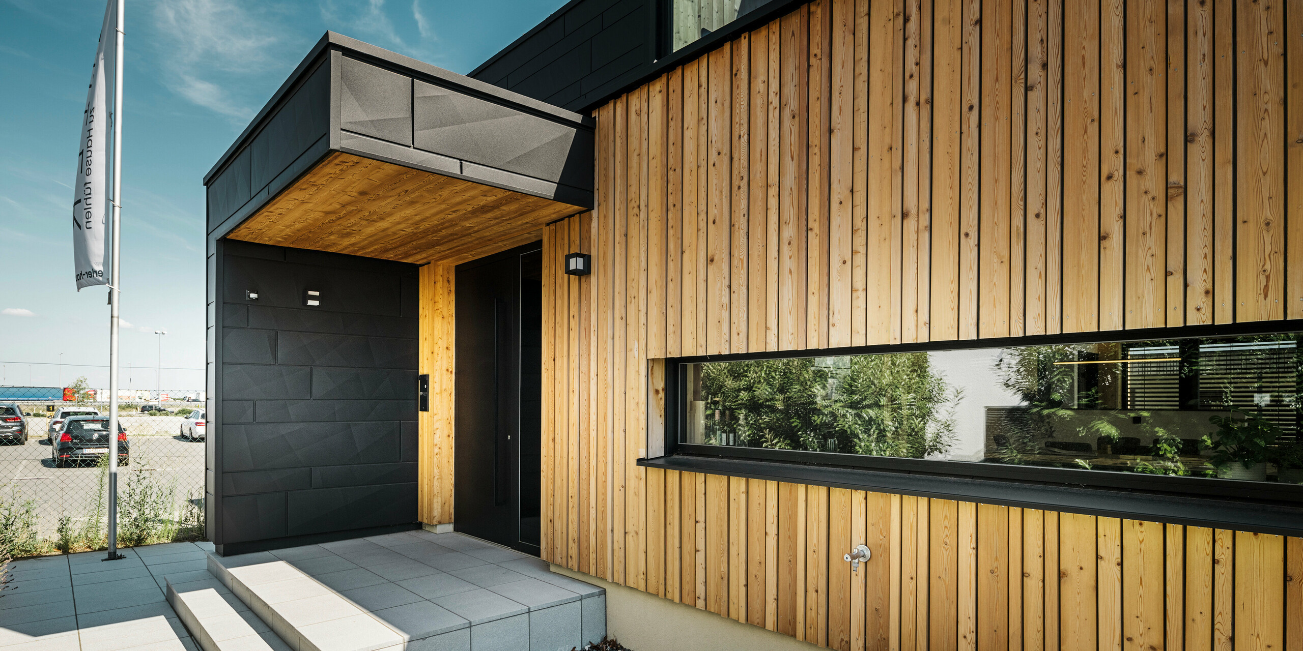 Ingresso laterale della casa campione Autark nel centro prefabbricati Blaue Lagune a Vösendorf, con una facciata moderna in legno naturale e  Doghe.X PREFA. L'immagine mostra la raffinata combinazione di rivestimento in legno e alluminio nel pregiato rivestimento P.10 nero.