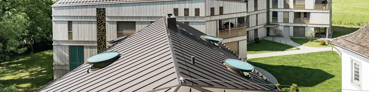 Moderno complesso residenziale a Liebwylen, in Svizzera, con un innovativo paesaggio di tetti in alluminio PREFALZ di colore marrone.  Il tetto con tecnologia a doppia aggraffatura integra numerosi lucernari rotondi e si inserisce armoniosamente nel paesaggio montano circostante. Sullo sfondo, un imponente paesaggio alpino e prati verdi.