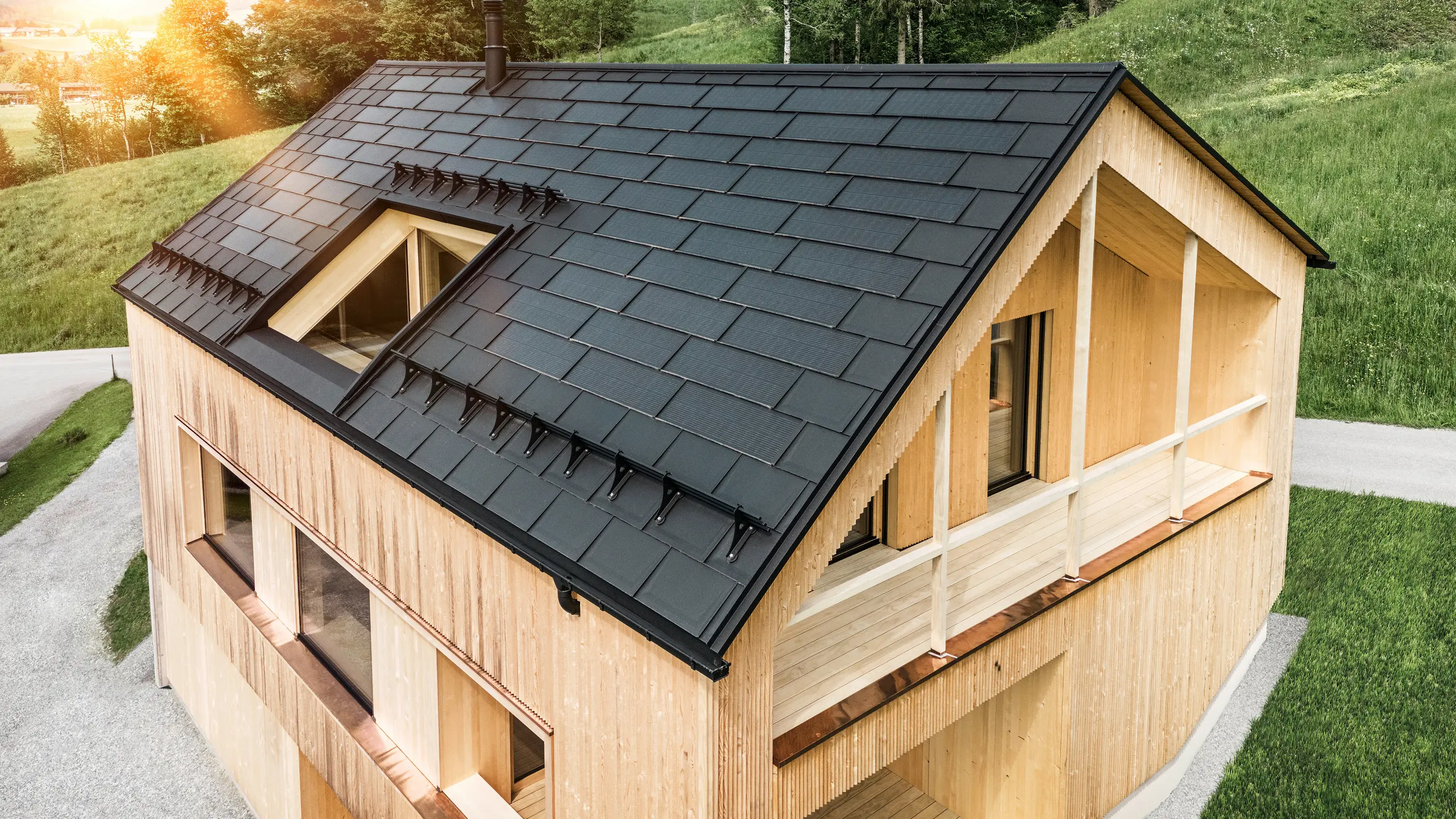 Vista di una moderna casa sostenibile in legno a Egg con un tetto PREFA Solar che si distingue per il suo aspetto scuro, strutturato ed elegante. Le tegole fotovoltaiche sono posizionati strategicamente trale classiche tegole del tetto R.16 per massimizzare il guadagno solare e allo stesso tempo armonizzarsi esteticamente con il design complessivo. La casa è in armonia con il verde circostante, mentre il caldo sole serale enfatizza la texture del legno naturale della facciata e sottolinea la solidità della costruzione. Un esempio armonioso di costruzione attenta all'ambiente e all'efficienza energetica.