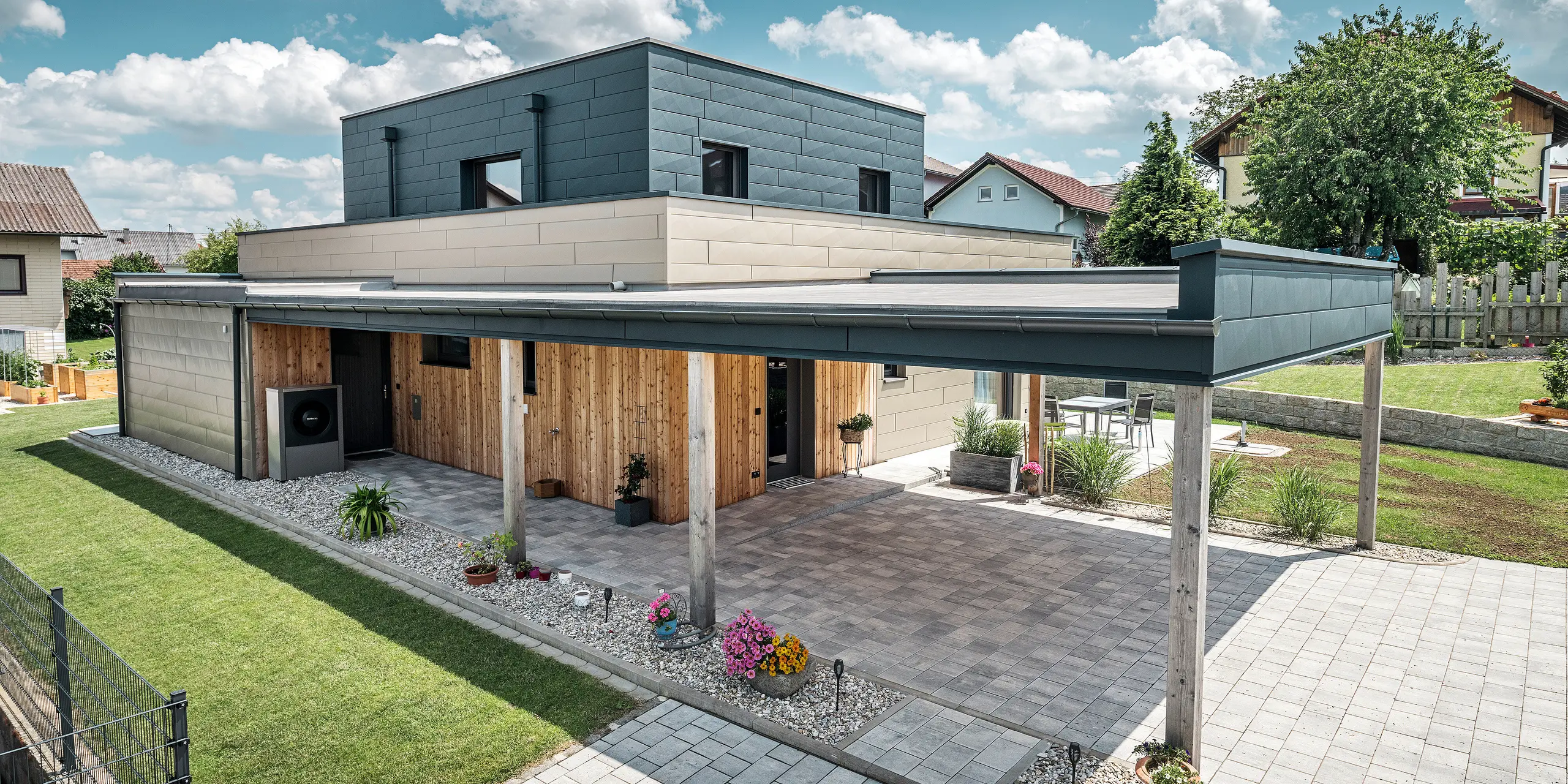 Vista aerea dell'ingresso di una moderna casa unifamiliare a Wippenham, in Austria, con una facciata bicolore in alluminio PREFA nei colori bronzo e antracite. La casa mostra l'elegante integrazione dei pannelli per facciate Siding.X e del rivestimento in legno. È chiaramente visibile il posto auto coperto, che si integra perfettamente nell'architettura della casa. Il giardino ben curato e la terrazza sottolineano il design moderno e funzionale dell'edificio.
