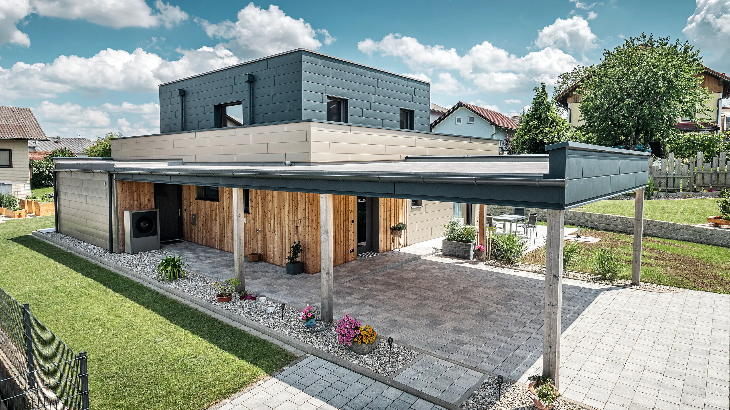 Vista aerea dell'ingresso di una moderna casa unifamiliare a Wippenham, in Austria, con una facciata bicolore in alluminio PREFA nei colori bronzo e antracite. La casa mostra l'elegante integrazione dei pannelli per facciate Siding.X e del rivestimento in legno. È chiaramente visibile il posto auto coperto, che si integra perfettamente nell'architettura della casa. Il giardino ben curato e la terrazza sottolineano il design moderno e funzionale dell'edificio.