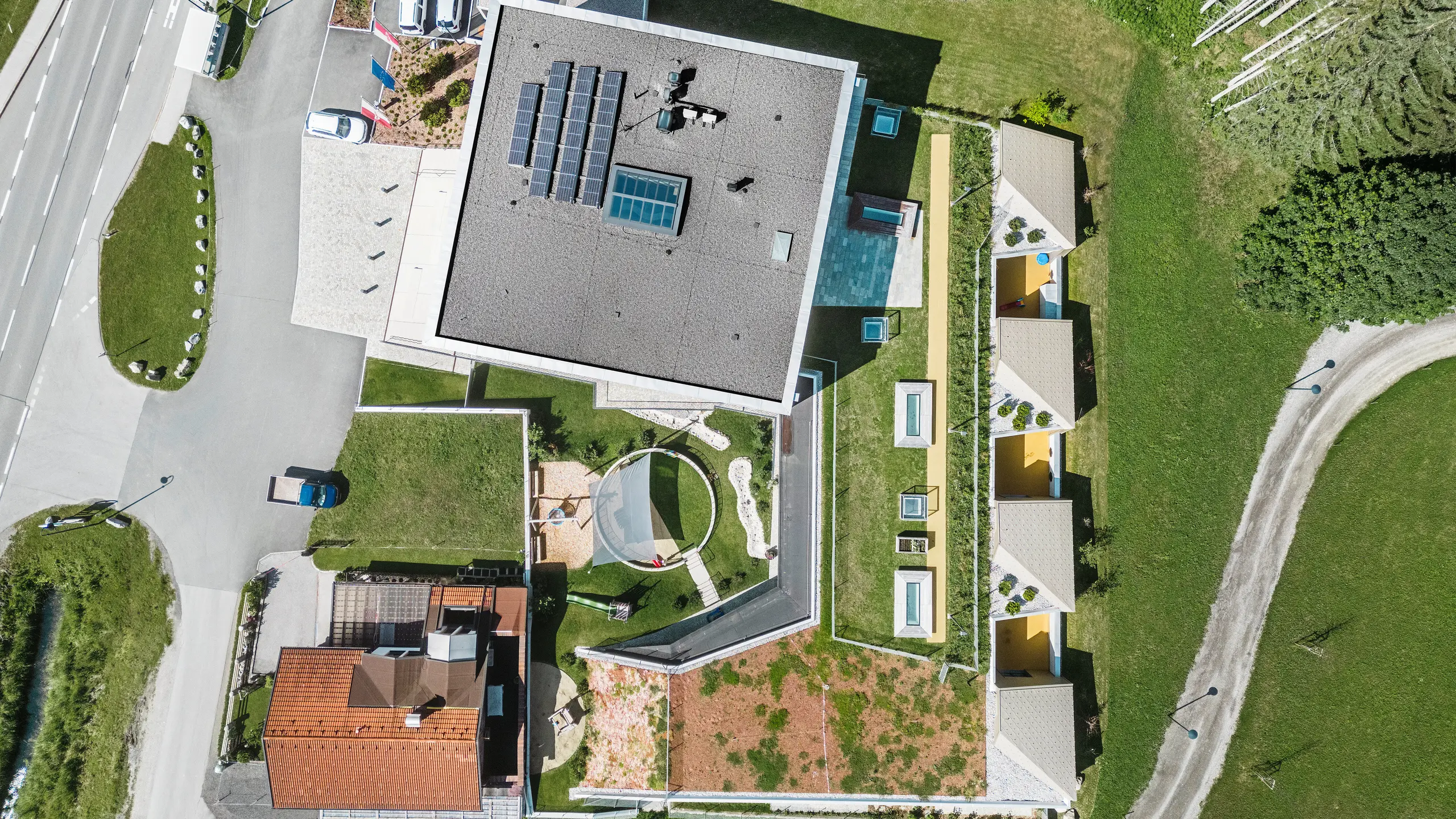Vista a volo d'uccello del centro educativo di Gerlos con un insieme di edifici moderni e tradizionali. L'edificio principale si presenta con un tetto grigio a falde dotato di pannelli solari ed è circondato da altri edifici con facciate gialle e tetti verdi. Sullo sfondo si vede il pittoresco paesaggio alpino boscoso, mentre in primo piano si vedono auto parcheggiate e una strada tortuosa.