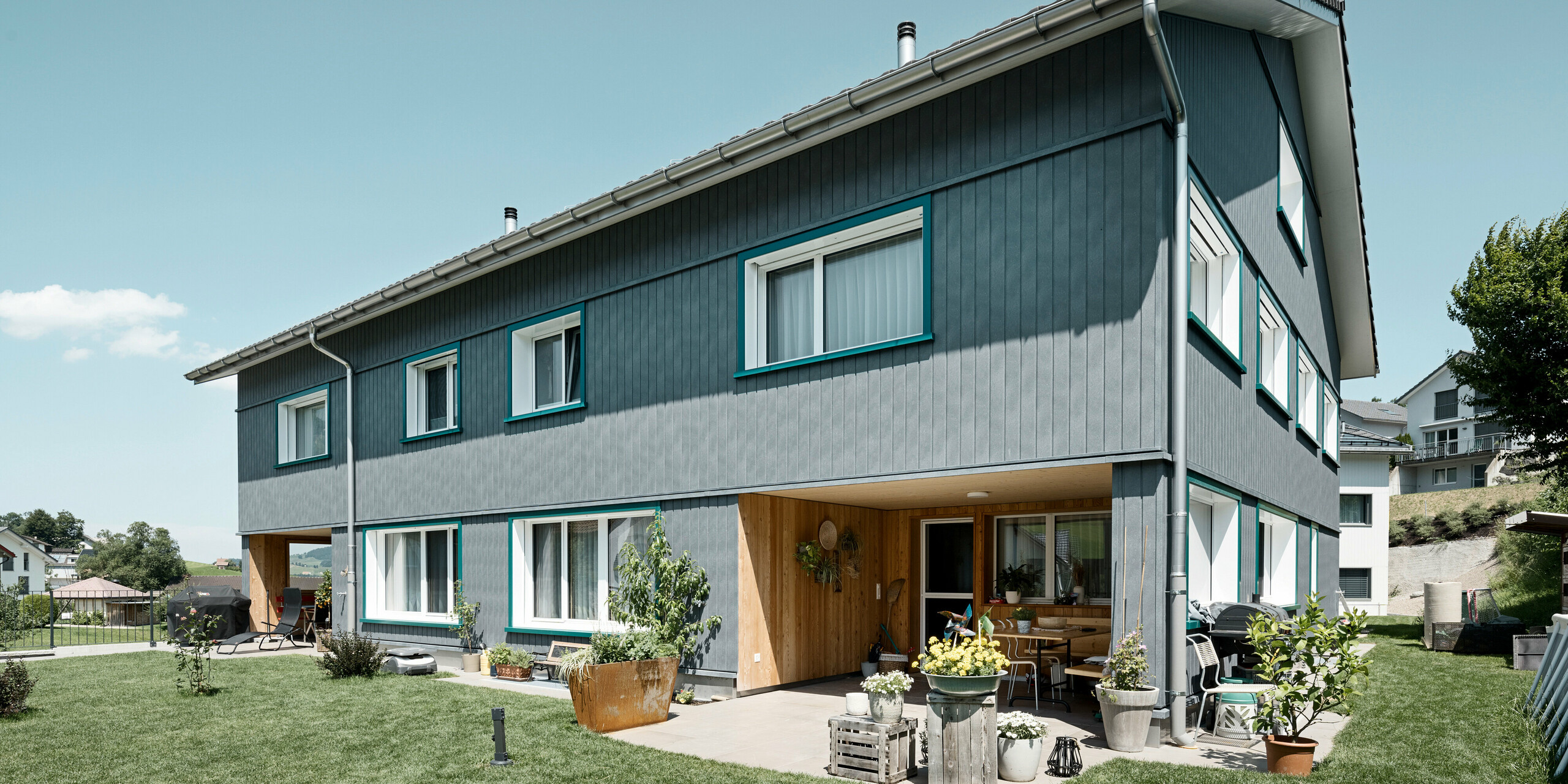 Gemütliches Mehrfamilienhaus in Weissbad, Schweiz, charakterisiert durch seine PREFA Siding Fassade in P.10 Steingrau, die sowohl Ästhetik als auch Langlebigkeit verspricht. Die vertikalen Linien der Sidings fügen sich nahtlos in die ländliche Umgebung ein und die lebhaften türkisen Fensterrahmen verleihen dem Gebäude eine einladende Note. Dieses Haus illustriert, wie PREFA Aluminiumprodukte die Balance zwischen Tradition und modernem Wohnkomfort meistern.