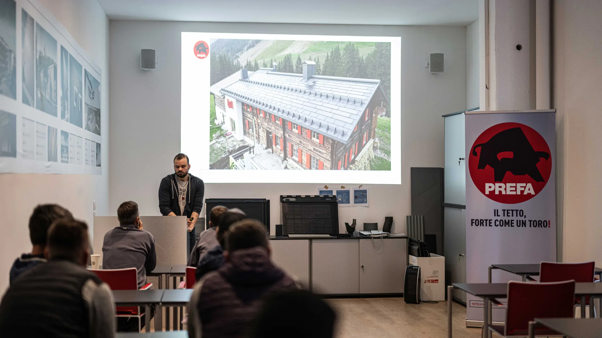 Ein Vortragender der PREFA Academy hält einen Vortrag über PREFA Dach- und Fassadenlösungen. Auf der Leinwand ist ein Bild eines Hauses mit PREFA Solardachplatte zu sehen. Die Teilnehmer sitzen im Schulungsraum und folgen der Präsentation. Im Raum ist ein PREFA Banner mit dem Slogan „Il tetto, forte come un toro!“ (Das Dach, stark wie ein Stier!) sichtbar.