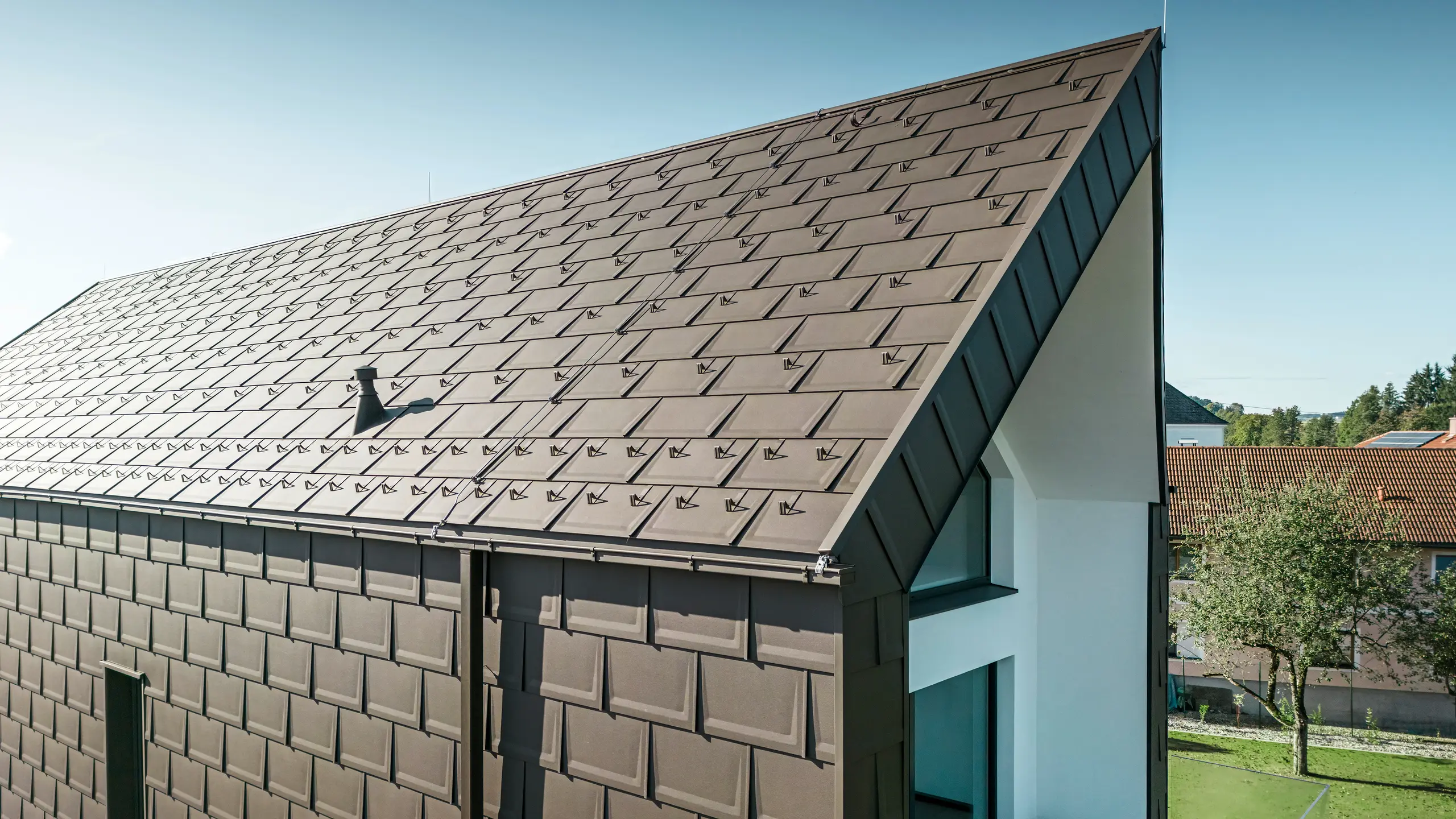Una vista prospettica del tetto e della facciata di una moderna casa unifamiliare a Neukirchen, in Austria. La tegola PREFA R.16 nella tonalità marrone P.10 offre una copertura esteticamente gradevole e funzionale, che si integra perfettamente nel design architettonico della casa. Il colmo affilato del tetto e la disposizione precisa delle tegole dimostrano la qualità di PREFA. I prodotti in alluminio di piccolo formato si integrano perfettamente nella facciata intonacata di bianco e nel paesaggio verde circostante.