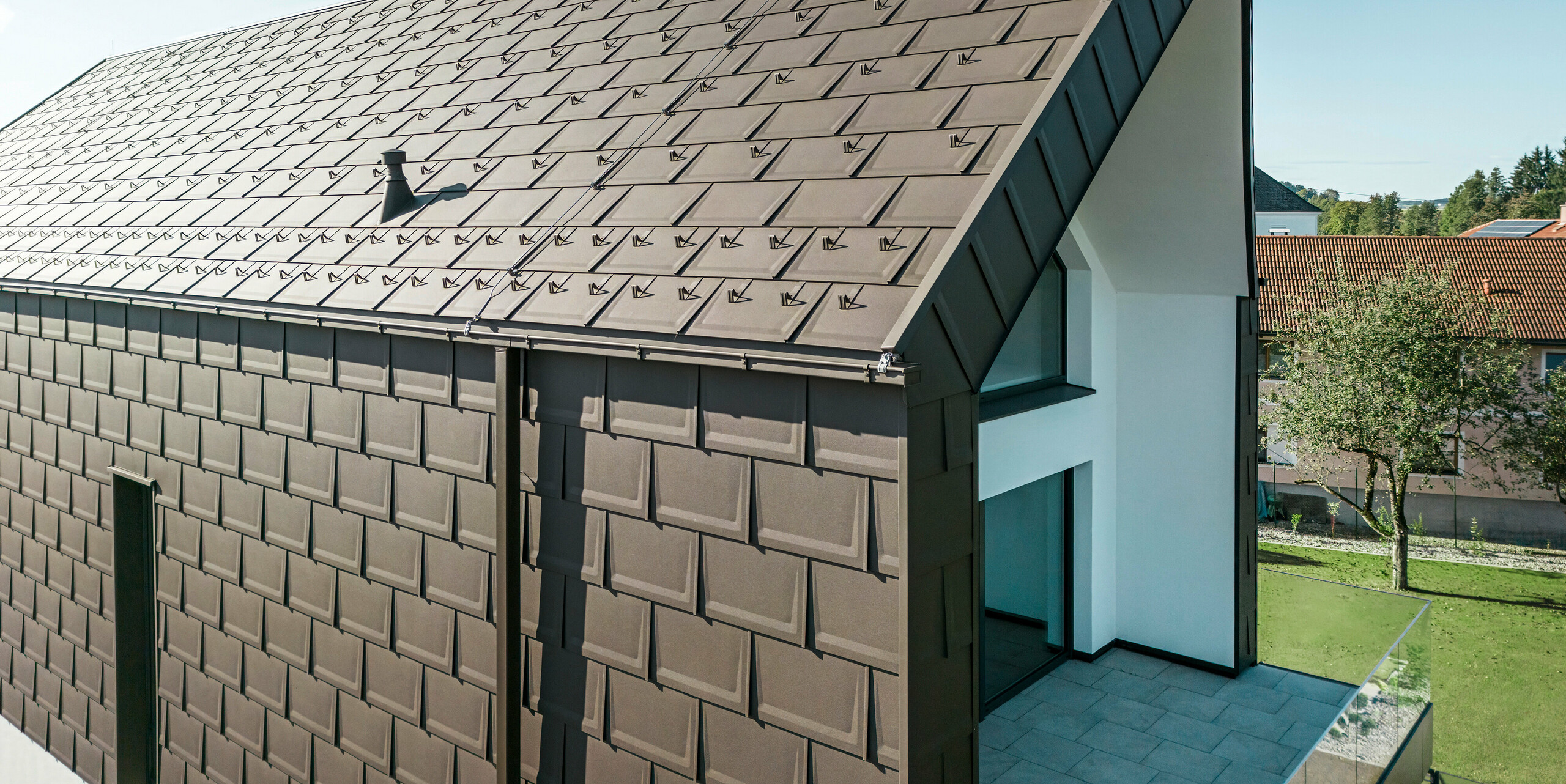 Perspektivische Ansicht des Daches und der Fassade eines modernen Einfamilienhauses in Neukirchen, Österreich. Die PREFA Dachplatte R.16 in P.10 Braun bietet eine ästhetische und funktionale Bedachung, die sich perfekt in die architektonische Gestaltung des Hauses einfügt. Der scharfe Dachfirst und die präzise Anordnung der Dachplatten demonstrieren die Qualität von PREFA. Die kleinformatigen Aluminiumprodukte harmonieren exzellent mit dem weiß verputzten Fassadenteil und der umliegenden grünen Landschaft.