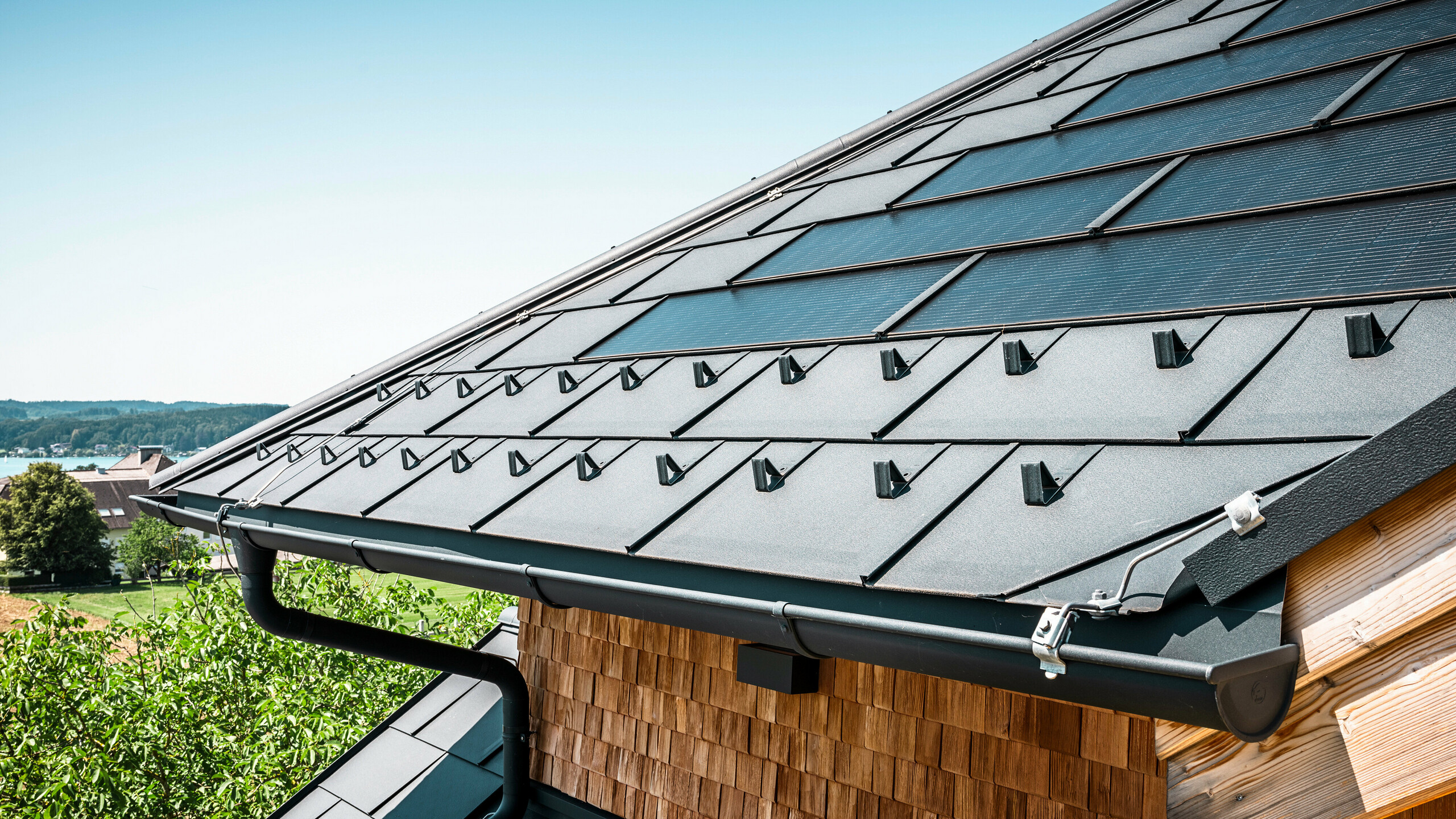 Die Zenz'n Stub'n in Schörfling am Attersee präsentiert ein neu saniertes Dach mit innovativen PREFA Solardachplatten und langlebigen PREFA Dachplatten R.16 in P.10 Anthrazit. Das stilvolle Aluminiumdach verbindet modernes Design mit nachhaltiger Energiegewinnung. Auch die Dachentwässerung, bestehend aus Ablaufrohr und Hängerinne in P.10 Anthrazit, fügt sich harmonisch in das Gesamtbild ein und unterstreicht die hochwertige Verarbeitung. Im Hintergrund blitzt das Blau des Attersees hervor.