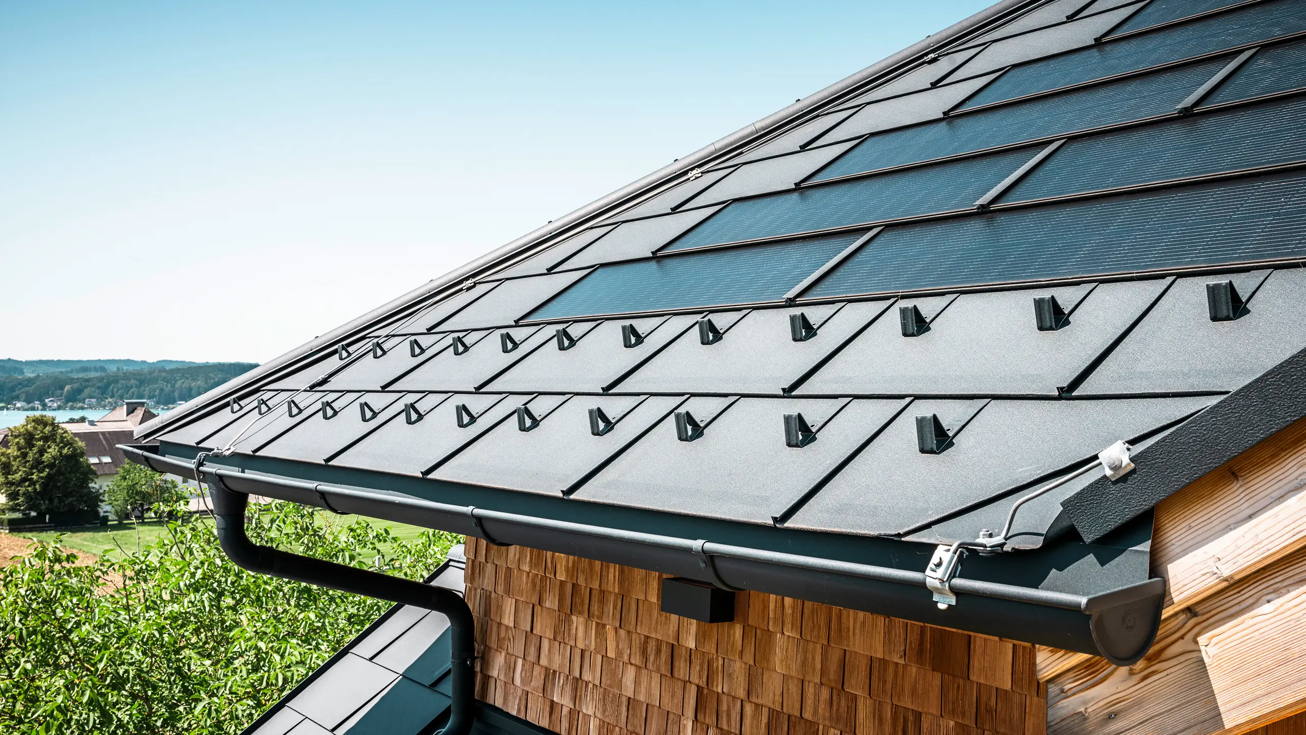 Die Zenz'n Stub'n in Schörfling am Attersee präsentiert ein neu saniertes Dach mit innovativen PREFA Solardachplatten und langlebigen PREFA Dachplatten R.16 in P.10 Anthrazit. Das stilvolle Aluminiumdach verbindet modernes Design mit nachhaltiger Energiegewinnung. Auch die Dachentwässerung, bestehend aus Ablaufrohr und Hängerinne in P.10 Anthrazit, fügt sich harmonisch in das Gesamtbild ein und unterstreicht die hochwertige Verarbeitung. Im Hintergrund blitzt das Blau des Attersees hervor.