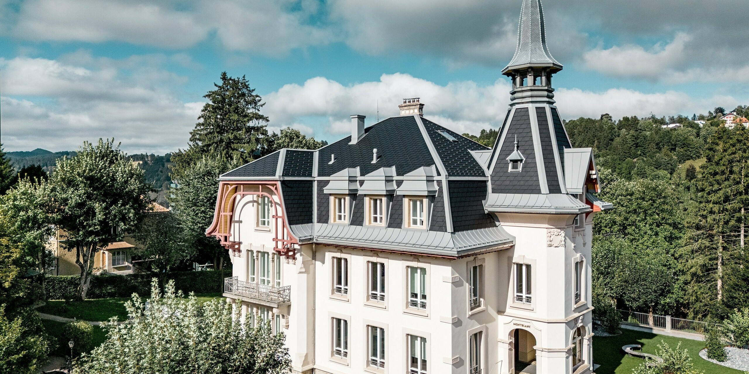 Atmosphärische Frontansicht der Manufacture Horlogère du Locle in der Schweiz mit eindrucksvollem Turm und dynamischer Dachlandschaft als Eyecatcher. Die komplexen, steil geneigten Dachflächen werden durch PREFA Dachschindeln in P.10 Schwarz präzise strukturiert, während PREFALZ in Patinagrau elegante Übergänge, Gauben und Rundungen formt. Das hochwertige Aluminiumdach überzeugt durch geringes Gewicht, hohe Widerstandsfähigkeit und extreme Langlebigkeit – eine perfekte Kombination aus architektonischer Ästhetik, technischer Präzision und nachhaltigem Schutz historischer Bausubstanz.