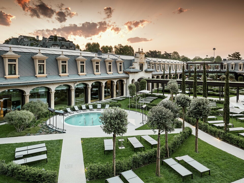 Abendstimmung im Thermalpark „De Montel – Terme Milano“ mit eindrucksvollem Blick auf die denkmalgeschützte Fassadenarchitektur im Jugendstil. Die Dacheindeckung mit PREFA Dachrauten 44 × 44 in P.10 Hellgrau verbindet Nachhaltigkeit mit Ästhetik. Im Vordergrund laden Liegewiesen, gepflegte Wege, Olivenbäume und ein rundes Thermalbecken zum Verweilen ein.