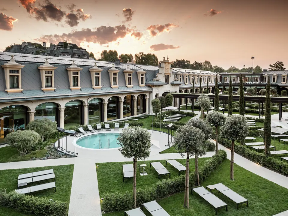 Abendstimmung im Thermalpark „De Montel – Terme Milano“ mit eindrucksvollem Blick auf die denkmalgeschützte Fassadenarchitektur im Jugendstil. Die Dacheindeckung mit PREFA Dachrauten 44 × 44 in P.10 Hellgrau verbindet Nachhaltigkeit mit Ästhetik. Im Vordergrund laden Liegewiesen, gepflegte Wege, Olivenbäume und ein rundes Thermalbecken zum Verweilen ein.