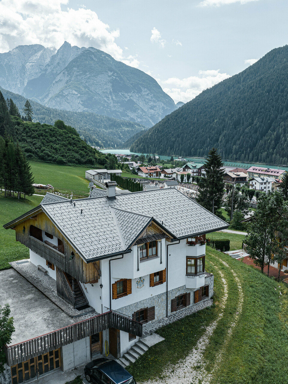 Luftaufnahme eines charmanten Bergchalets in Auronzo di Cadore, Italien, nach der Dachsanierung mit PREFA Aluminiumziegeln in Hellgrau. Das Chalet erhebt sich auf einer grünen Wiese mit einer beeindruckenden Bergkette im Hintergrund und zeigt ein malerisches Zusammenspiel von modernisierten Bauelementen und traditioneller Alpenarchitektur. Über dem Eingangsbereich ist eine Spitzgaube mit einem Holzgiebel zu sehen.
